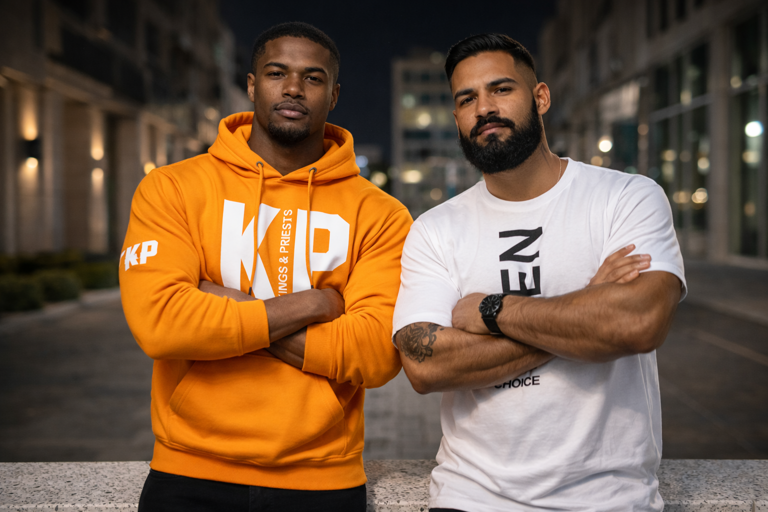 Two men standing with crossed arms outdoors at night in an urban area, one wearing an orange hoodie and the other a white t-shirt.
