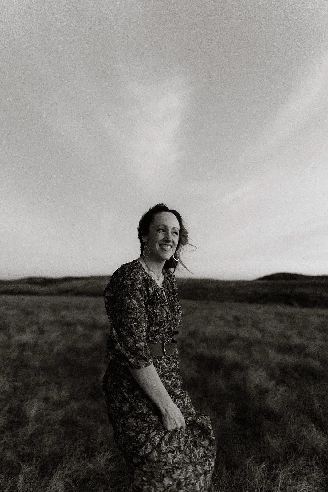 A woman standing in a grassy field, smiling, in black and white photo with open sky above.