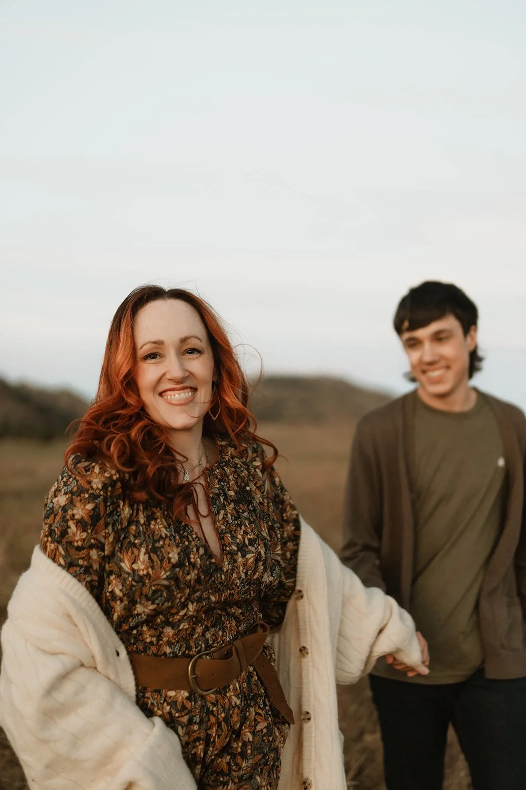 Smiling woman with red wavy hair in floral dress and cardigan holding hands with smiling man in an outdoor landscape at sunset.