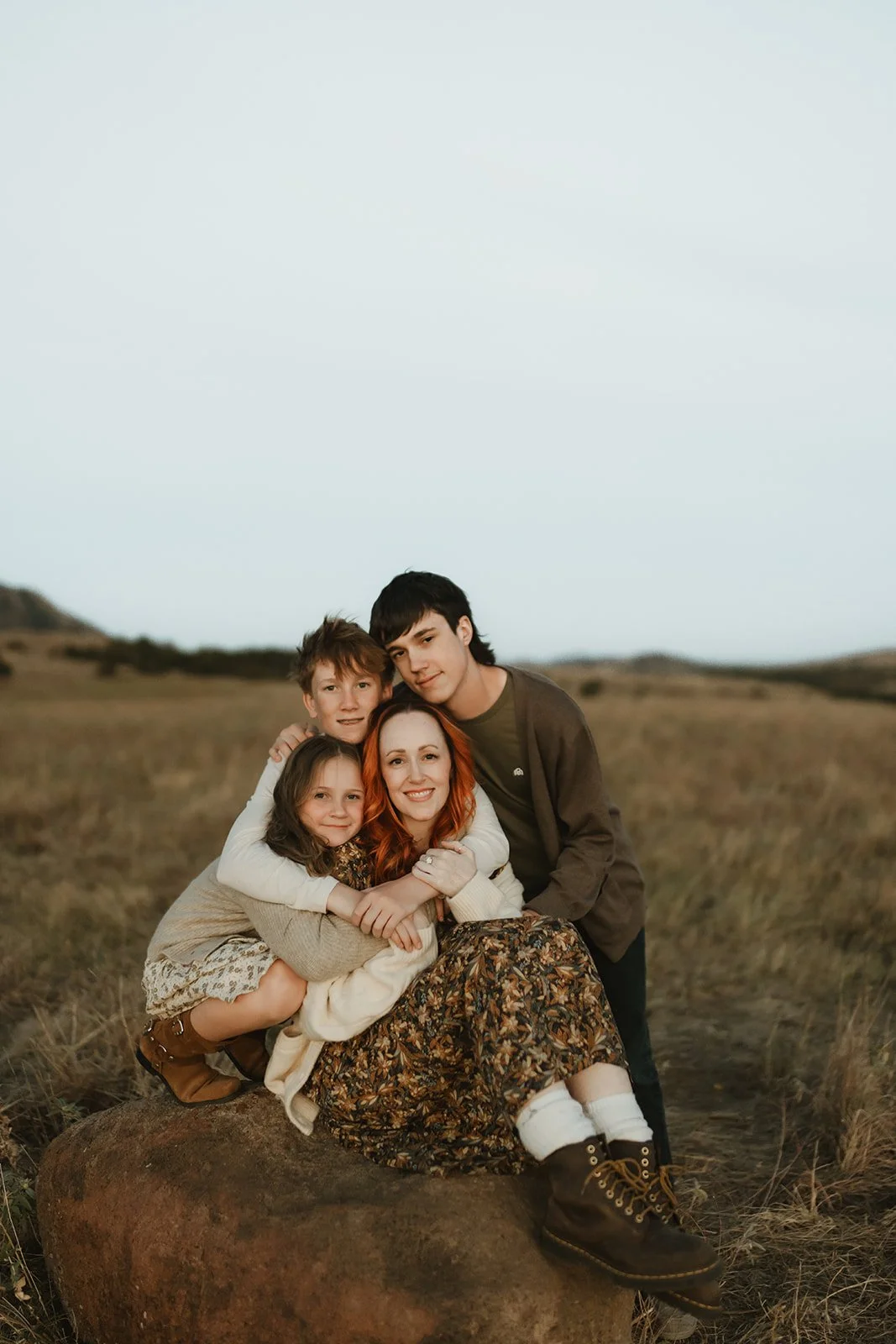 A woman with red hair and three children sitting on a large rock in an open grassy field during daylight.