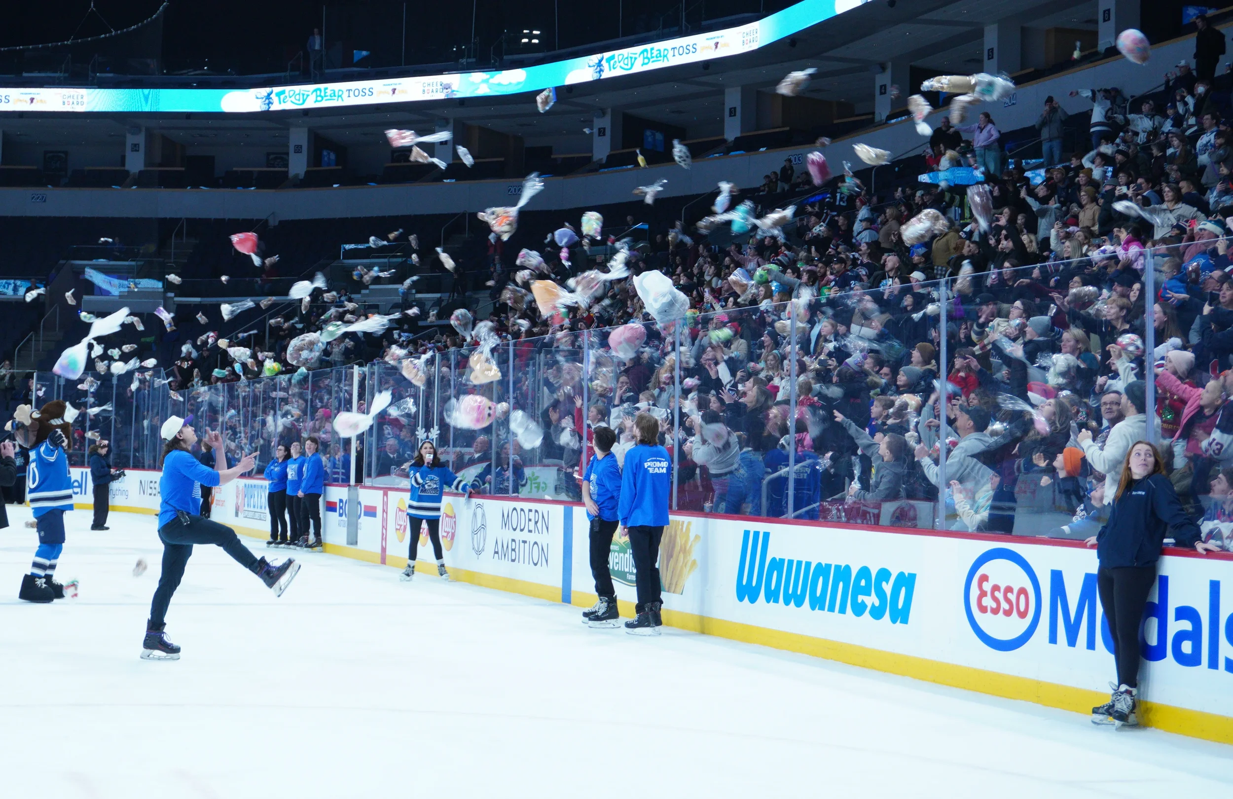 Ice hockey arena filled with fans tossing stuffed toys onto the ice during a charity event, with players and officials along the rink.