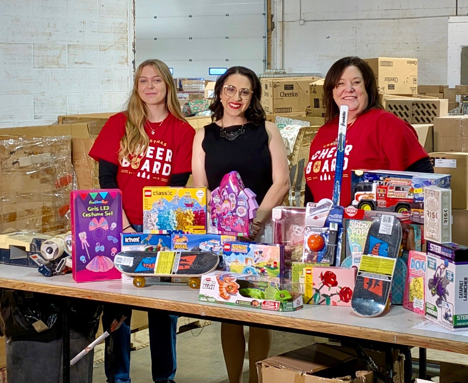 Three women standing behind a table filled with toys and gift items in a warehouse setting, some wearing red "Christmas Cheer Board" shirts, smiling.