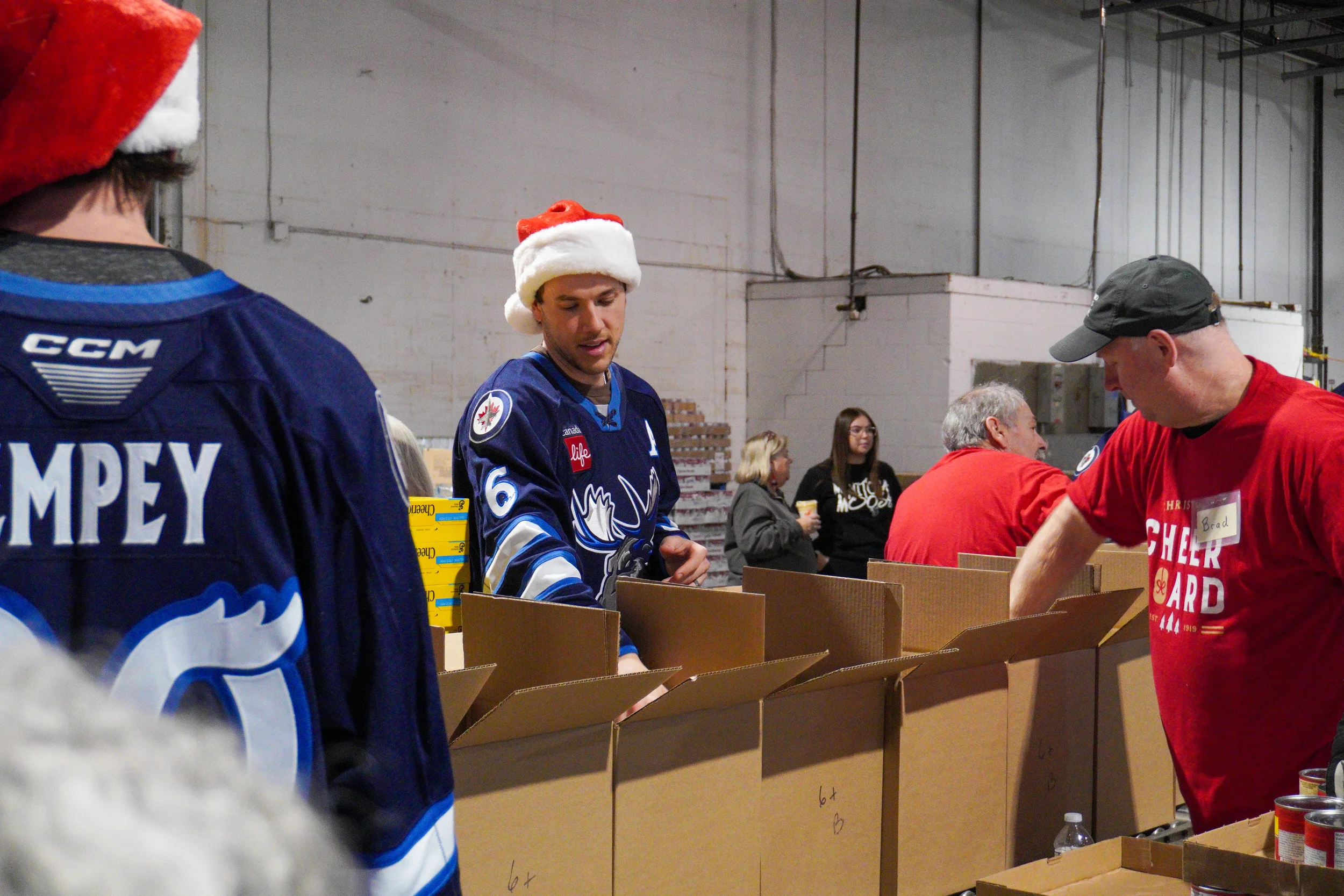 People packing food donations at a table in a warehouse, some wearing Manitoba Moose jerseys and red Santa hats.