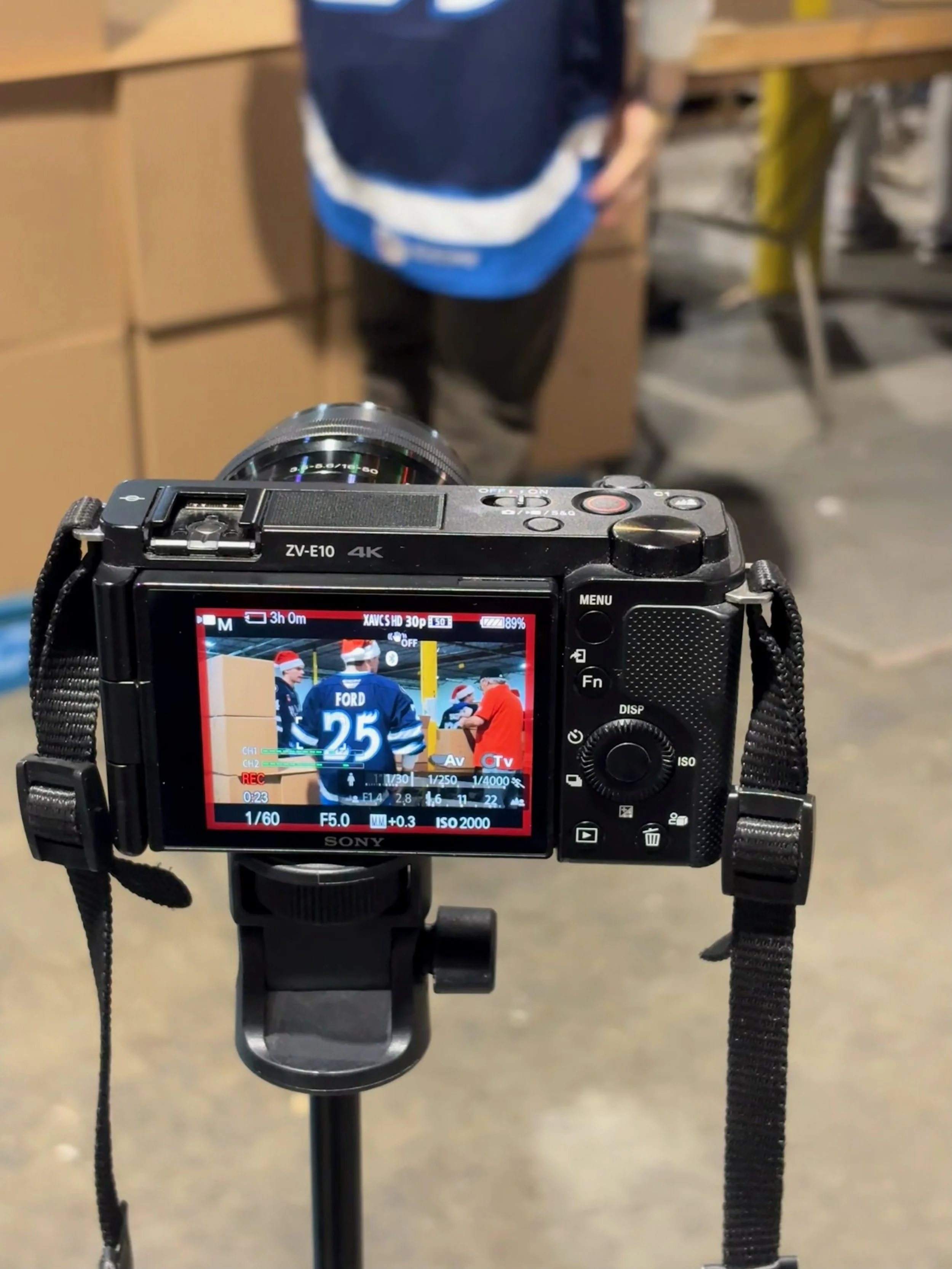 Digital camera mounted on tripod capturing an image of people wearing hockey jerseys and Santa hats, visible on the camera's screen.