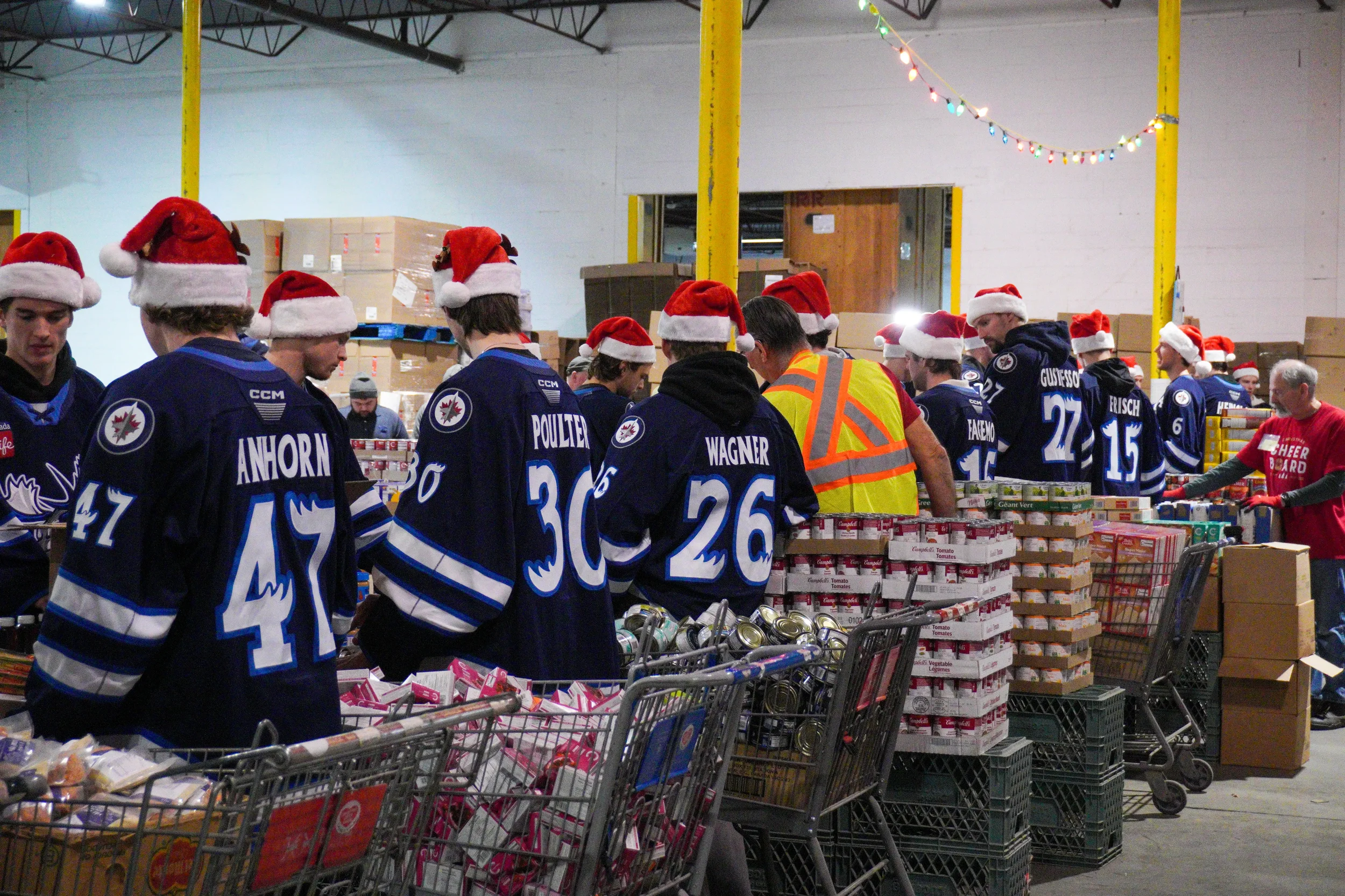 Group of volunteers wearing hockey jerseys and Santa hats shopping for food at a distribution center with Christmas lights hanging overhead.