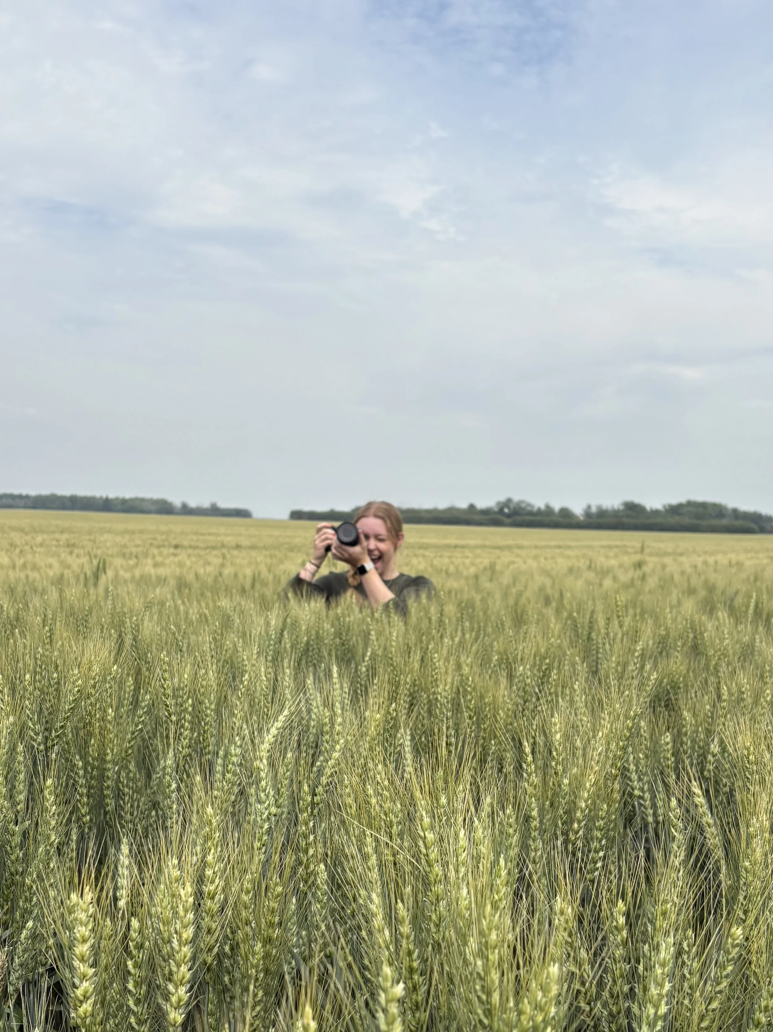 Person smiling and taking a photo with a camera in a green wheat field under a cloudy sky.