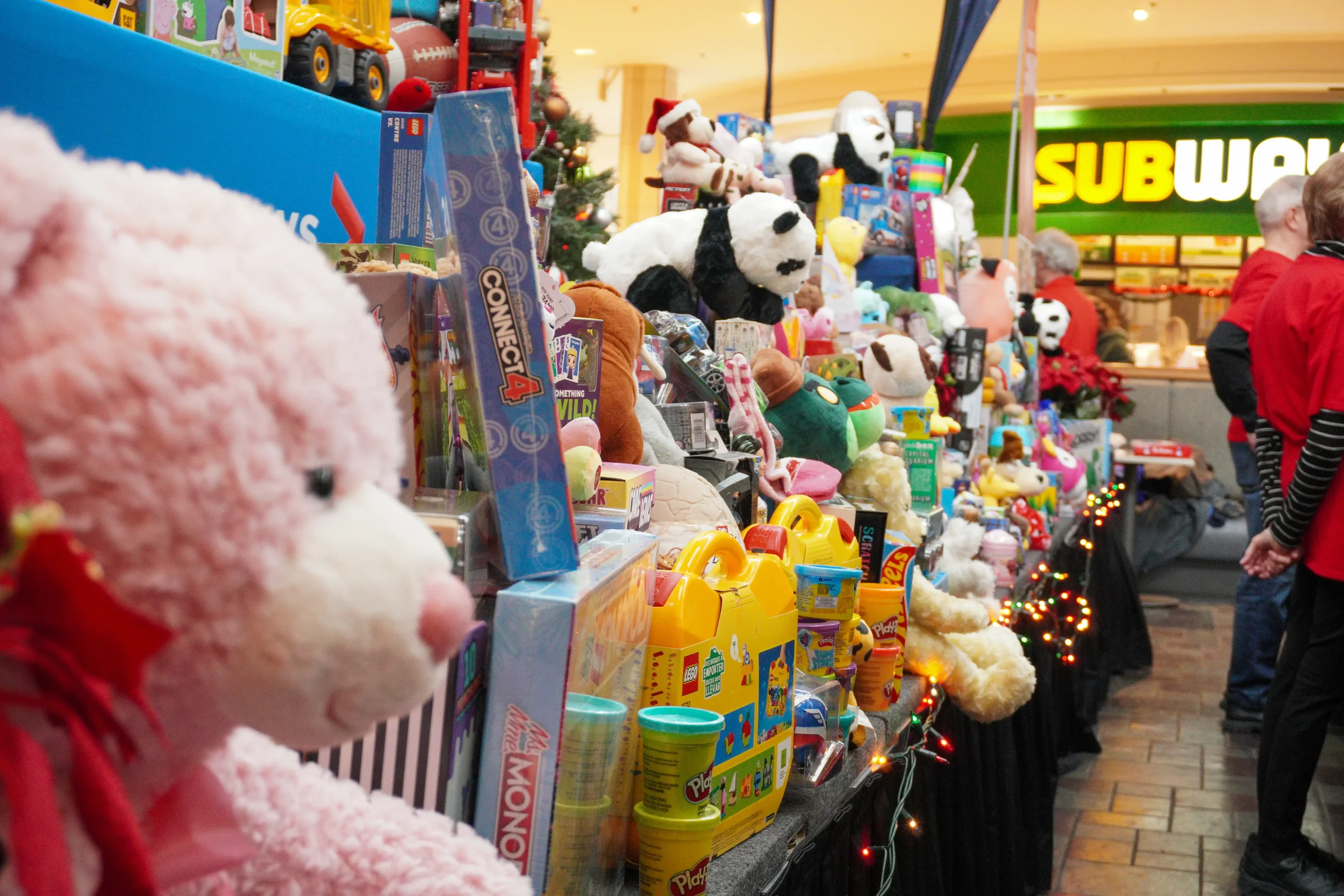 A display of Christmas plush toys and toys at a store, with people shopping and a Subway restaurant in the background.
