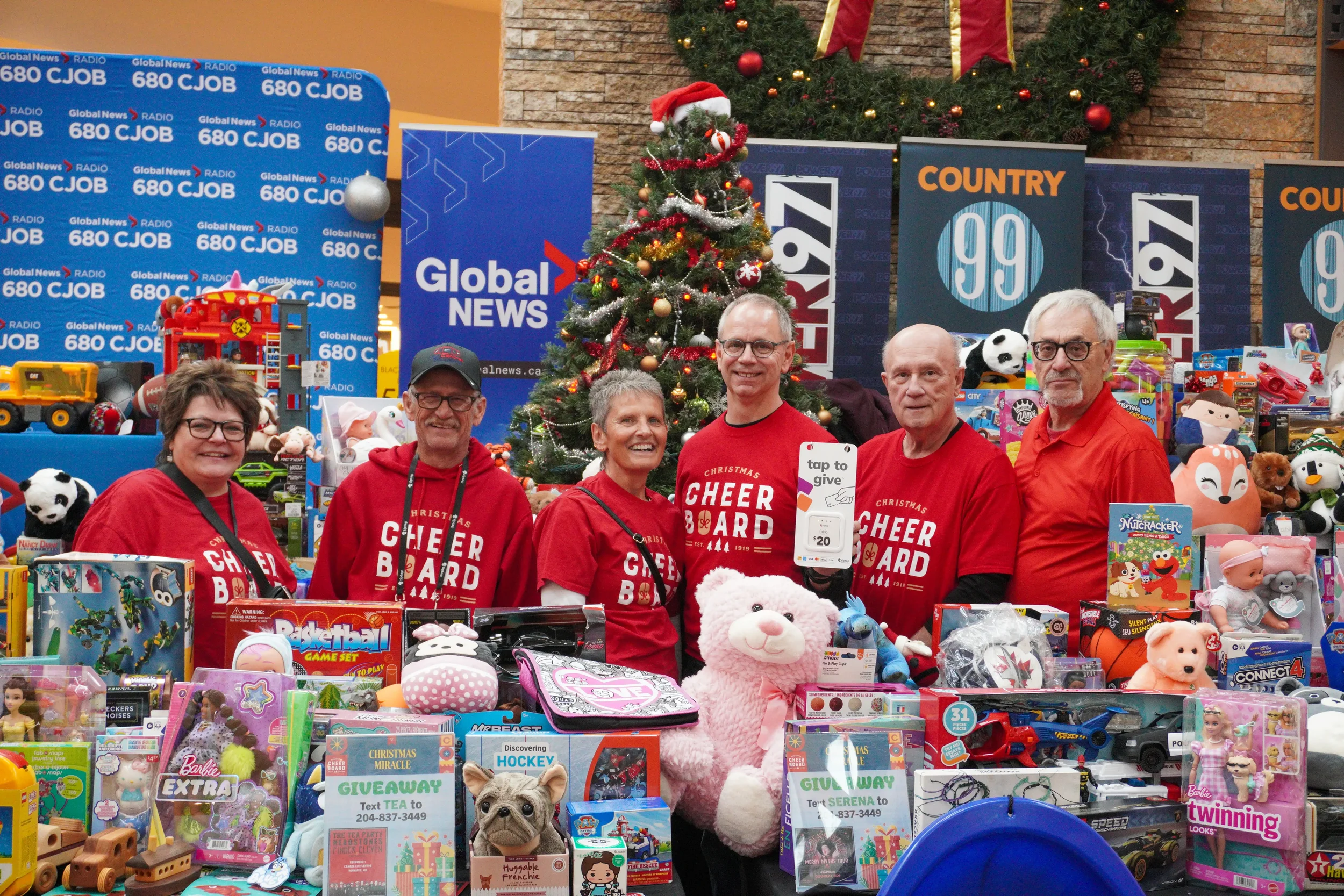 Six people standing behind a table filled with toys, with a decorated Christmas tree in the background, at a toy drive event organized by Global News and CJOB.