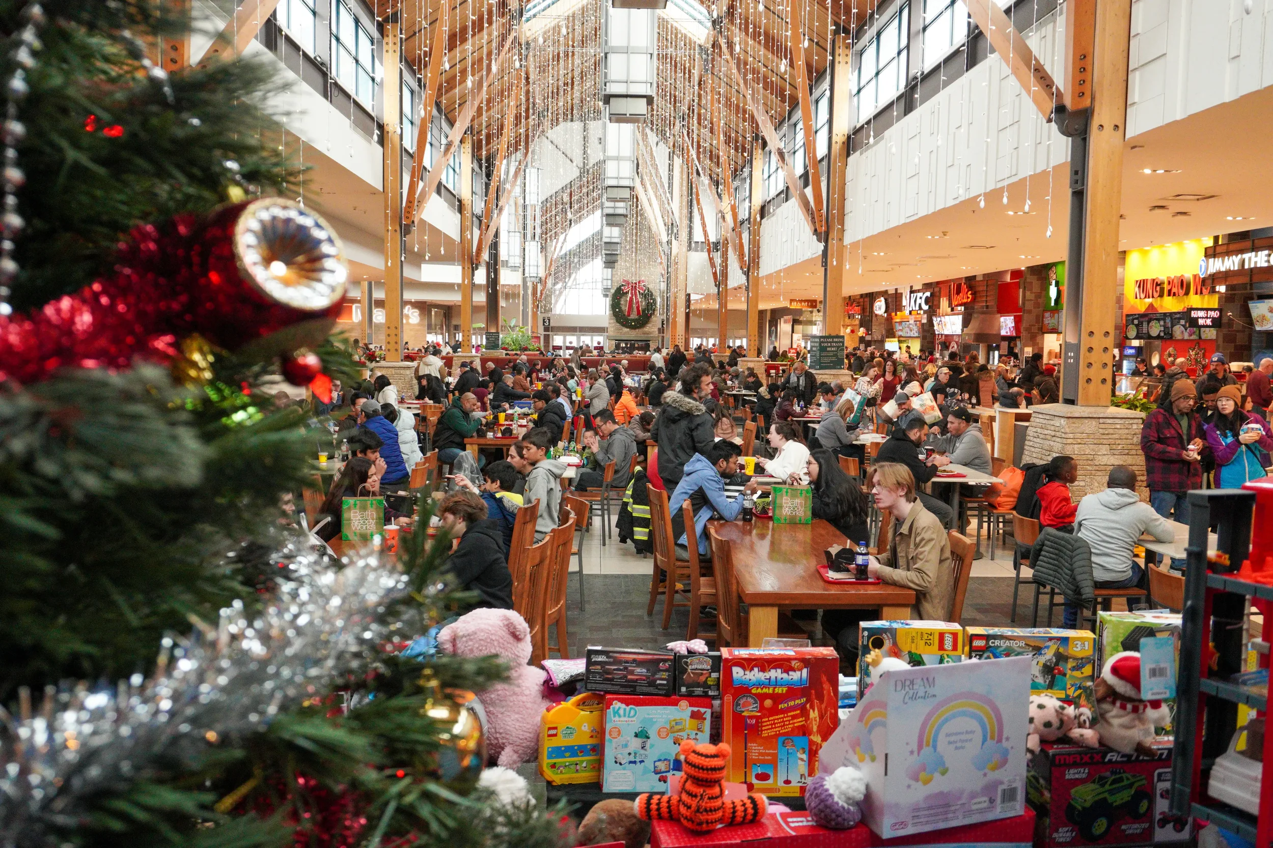 A busy shopping mall decorated for Christmas with a decorated Christmas tree in the foreground and toys on display. The mall has a high arched ceiling with holiday lights and a large wreath hanging in the center. Many people are sitting and dining at