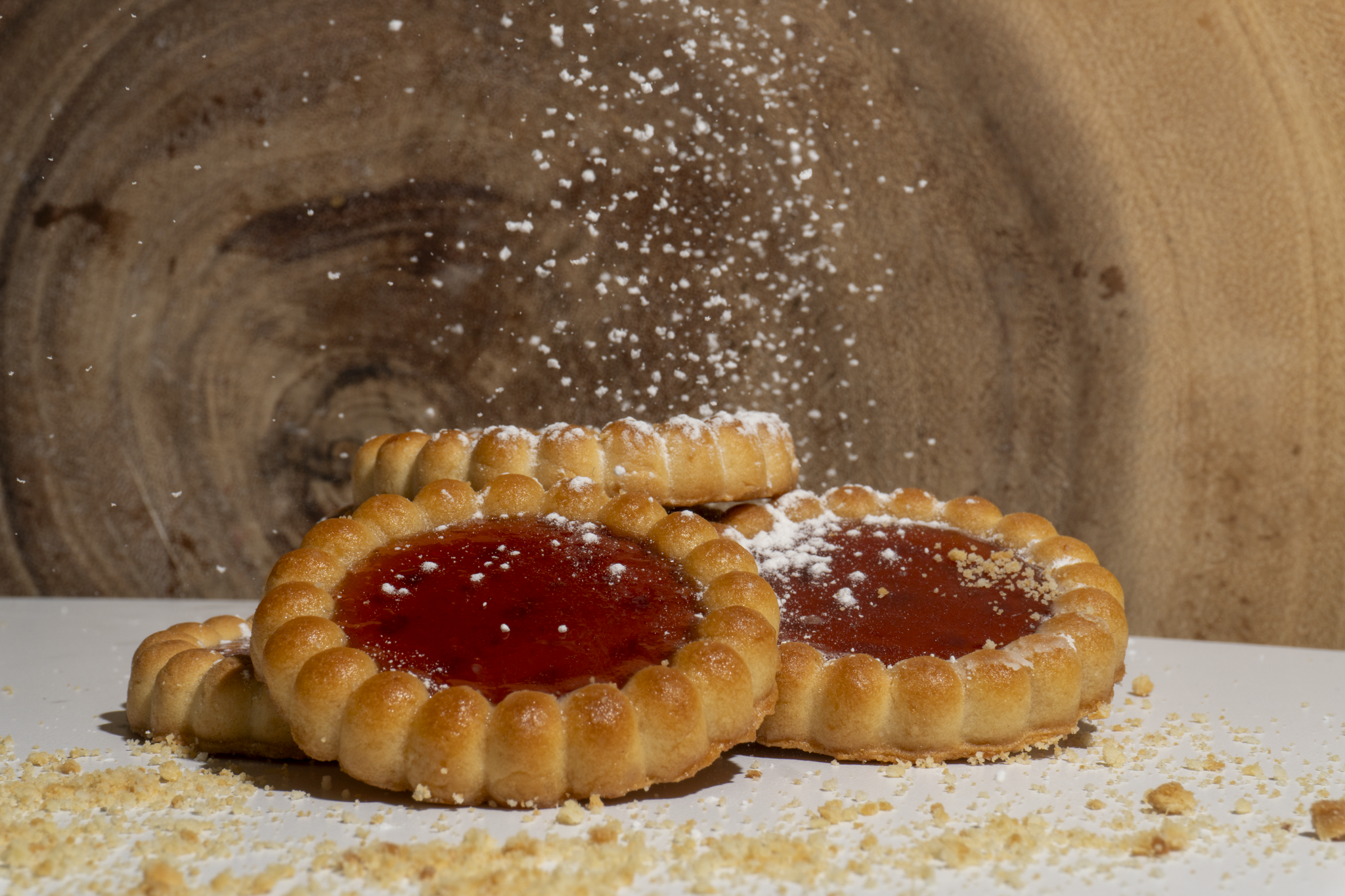 Three fruit tarts with red jelly and flaky crust, sprinkled with powdered sugar, on a white surface with crumbs and a wooden background.
