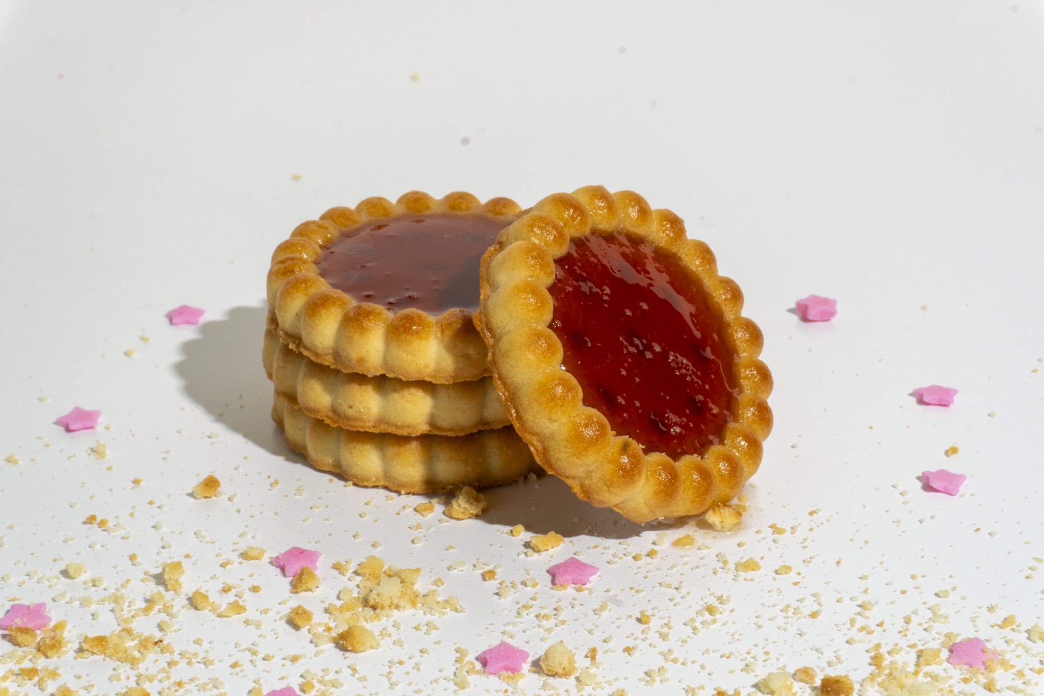 Four golden cookies with red jam filling, stacked on a white surface decorated with pink flower-shaped sprinkles and crumbs.
