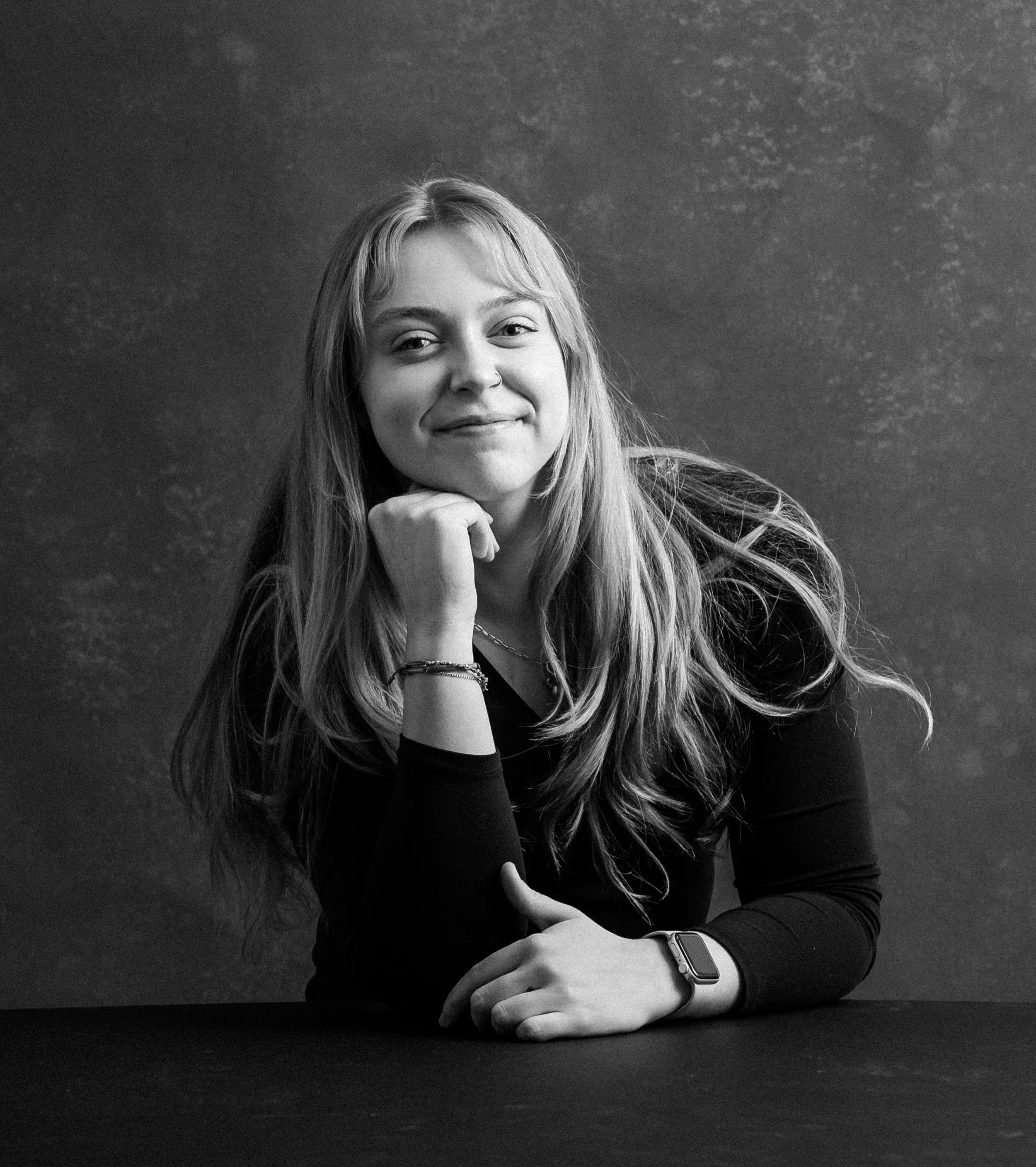 A young woman with long hair is resting her chin on her hand, smiling slightly. She's wearing a dark long sleeve shirt and sits at a table in front of a textured dark background.
