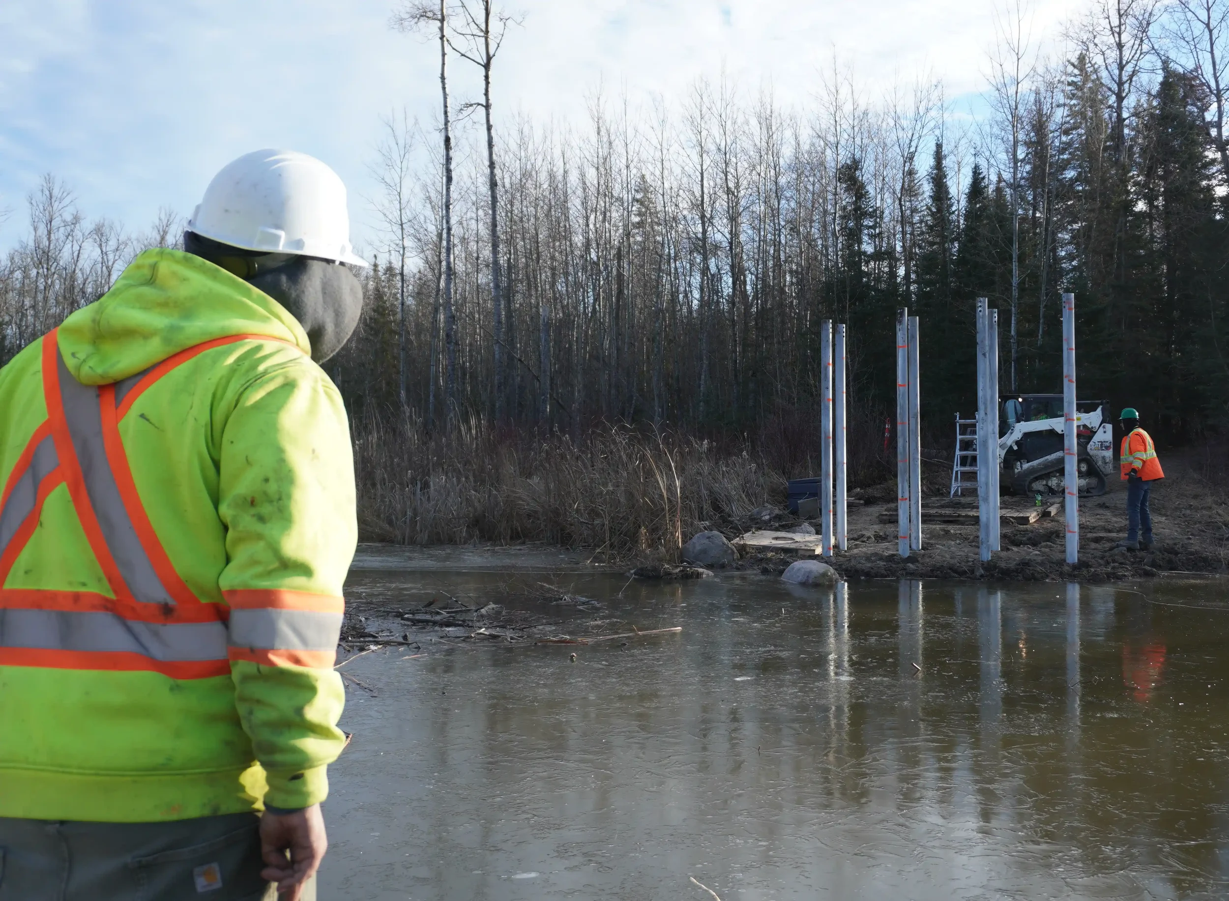 Construction workers working near a river, installing structural poles with equipment and trees in the background.