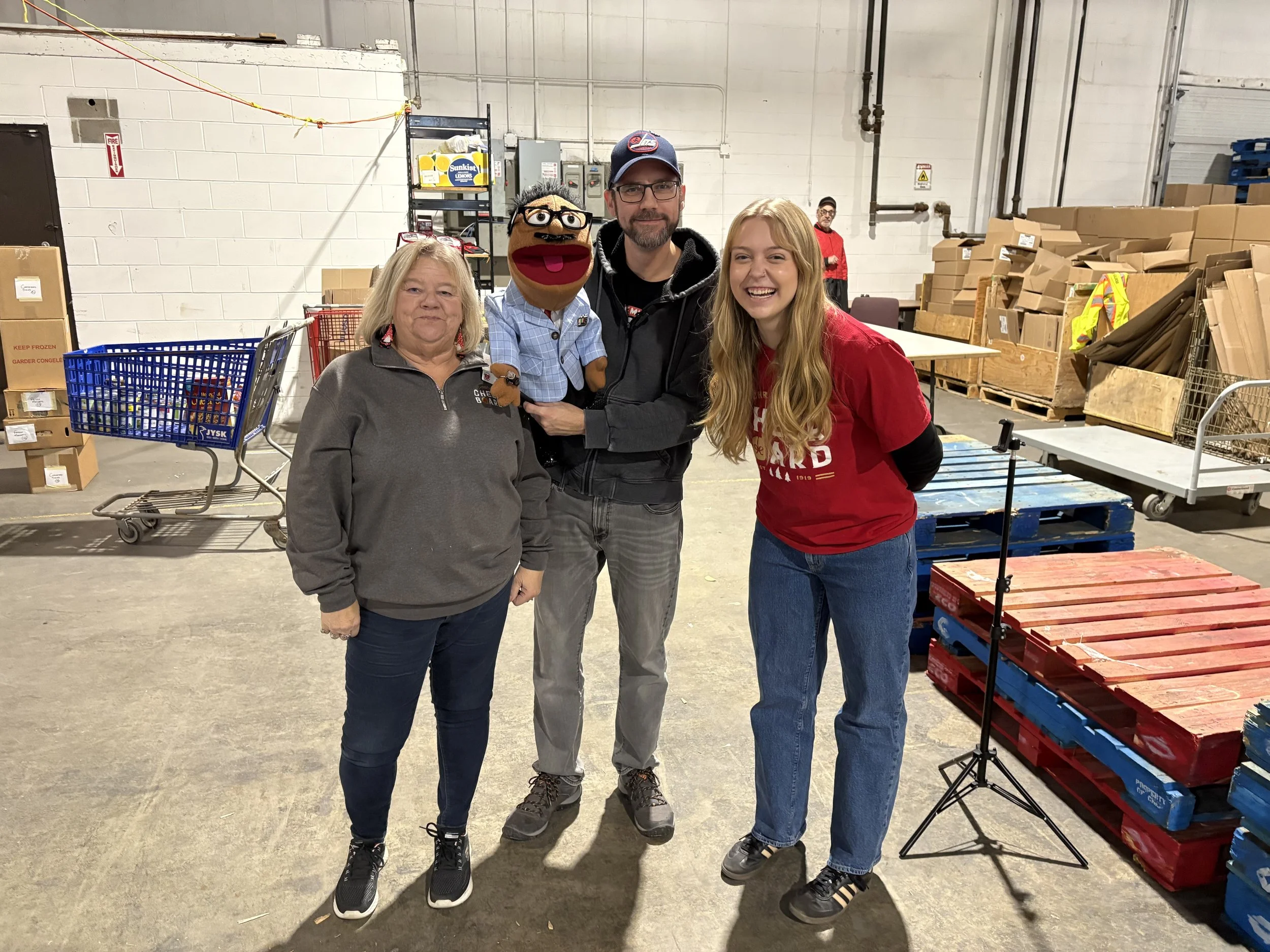 Three women and a man with a puppet in a warehouse, with boxes and carts in the background.