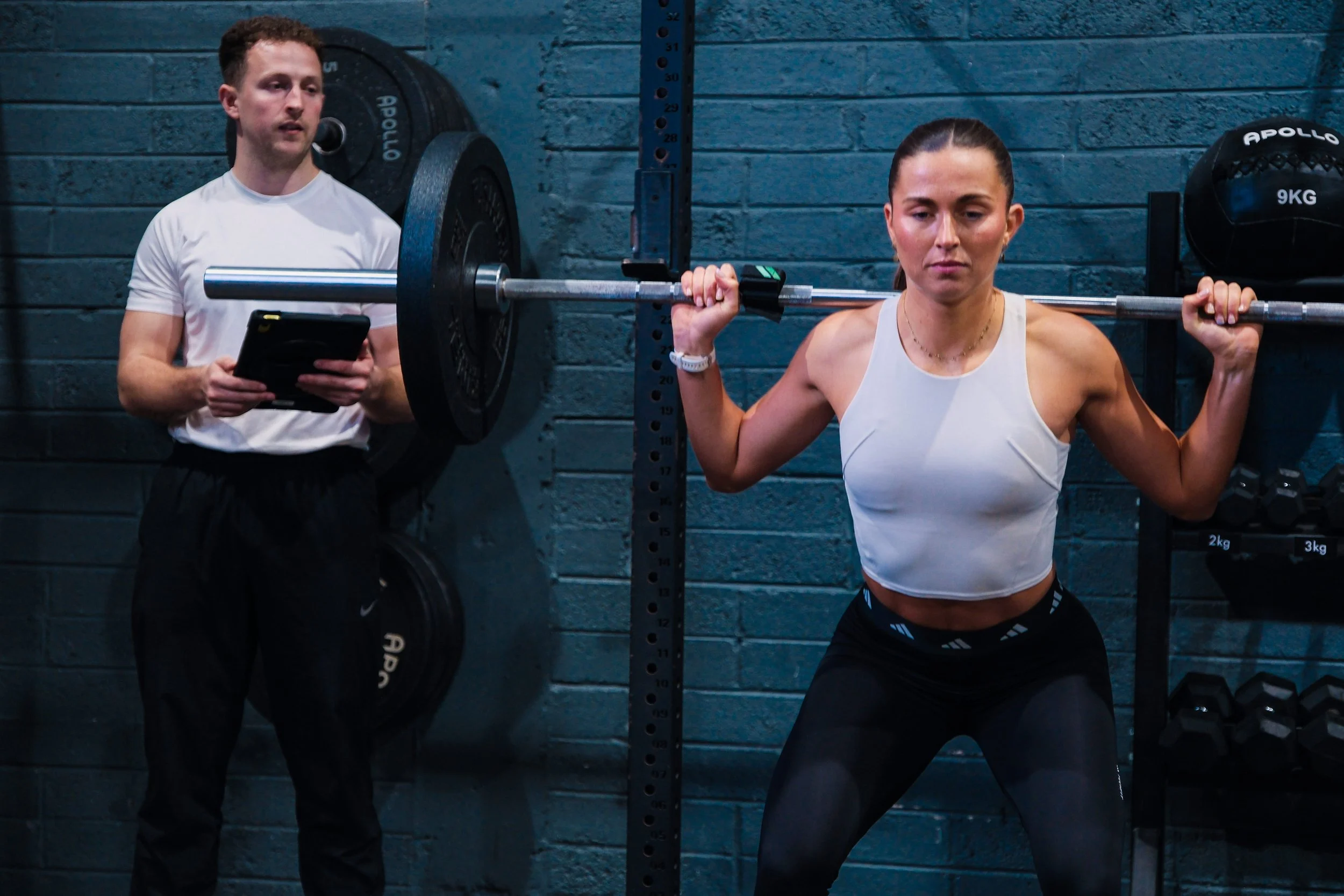 A woman with dark hair tied back is lifting a barbell on her shoulders during a workout, while a trainer in a white shirt is observing and holding a clipboard in a gym with blue brick walls.
