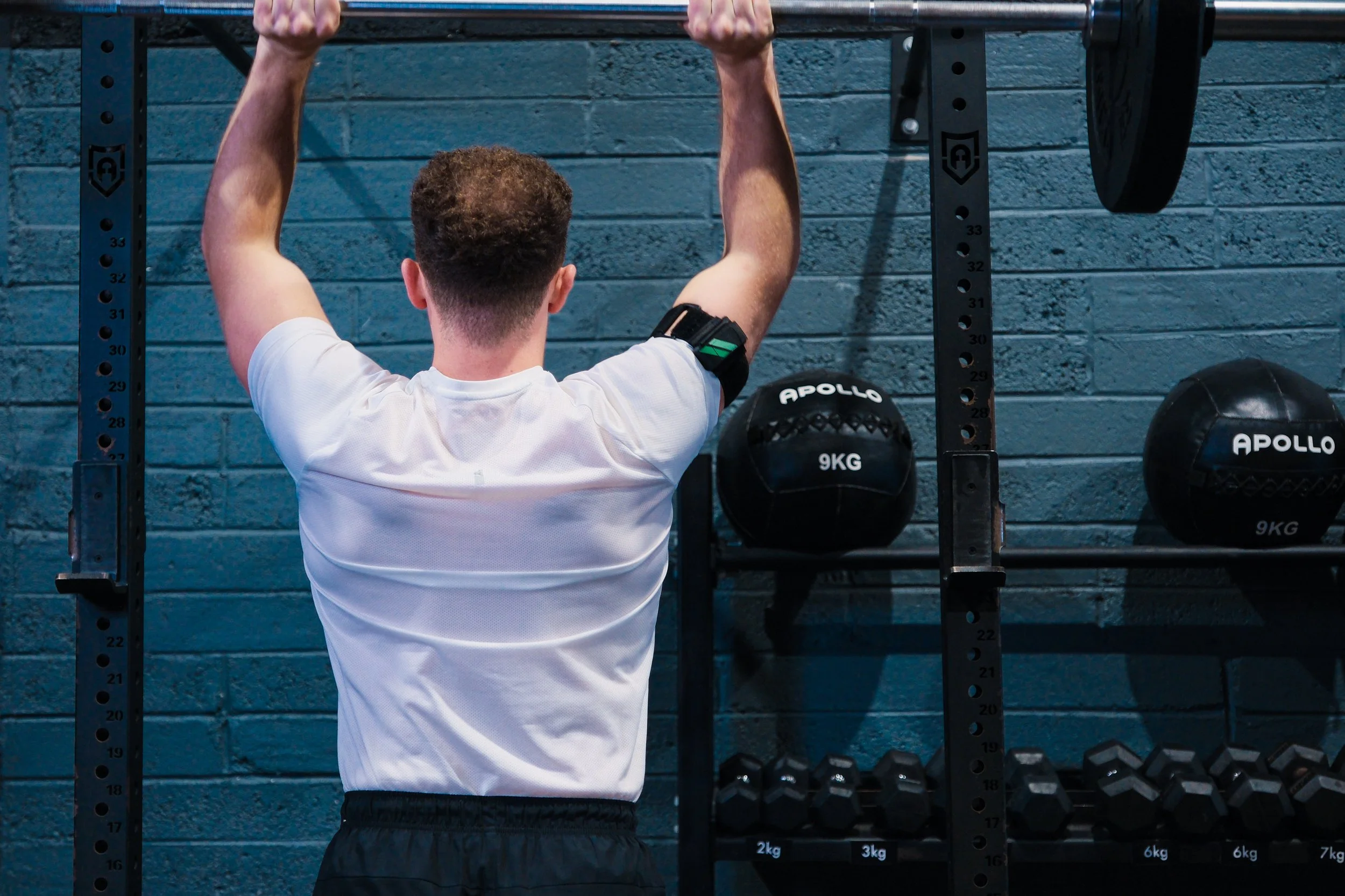 A man seen from behind, wearing a white t-shirt and black shorts, lifting a barbell overhead in a gym. The gym has a blue brick wall, a black metal squat rack, and black weight plates labeled 'APOLLO' hanging on the sides.