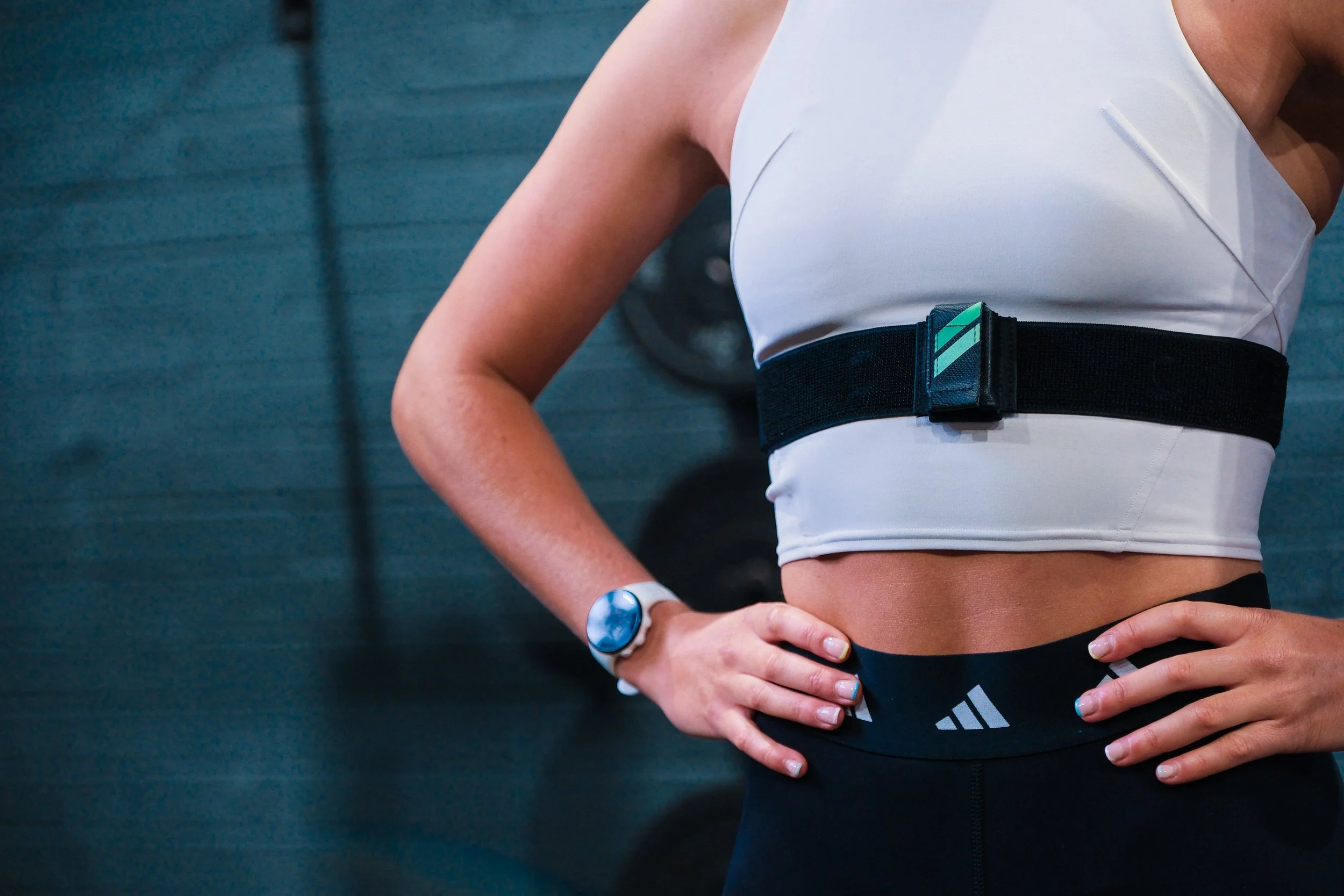 Close-up of a woman with athletic build wearing a white sports top, black workout leggings, and a black waistband with a small logo, standing with hands on hips in a gym setting.