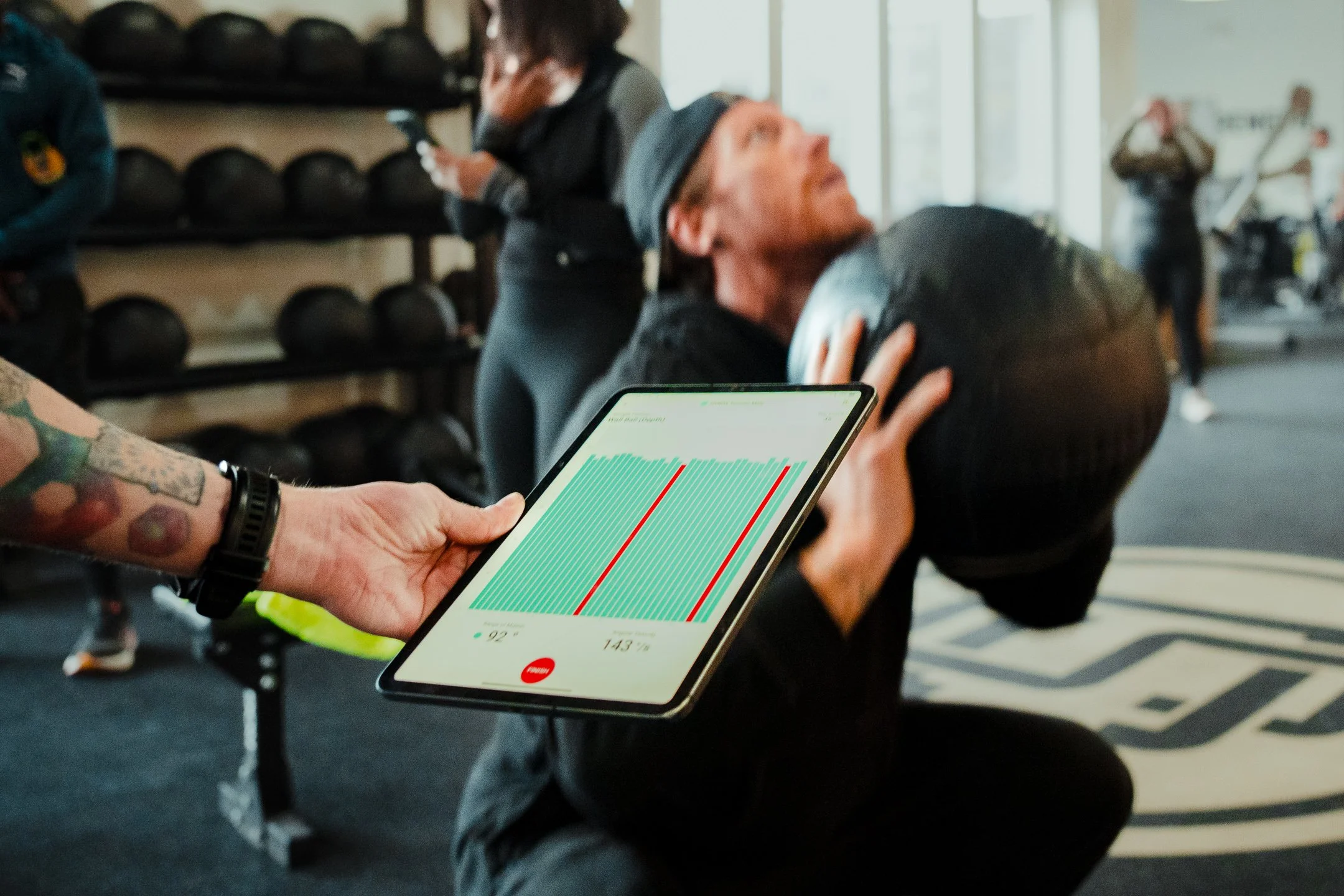 A person with tattoos on their arm holding a tablet displaying a running app with heart rate and pace information, seated in a gym with other people in the background.