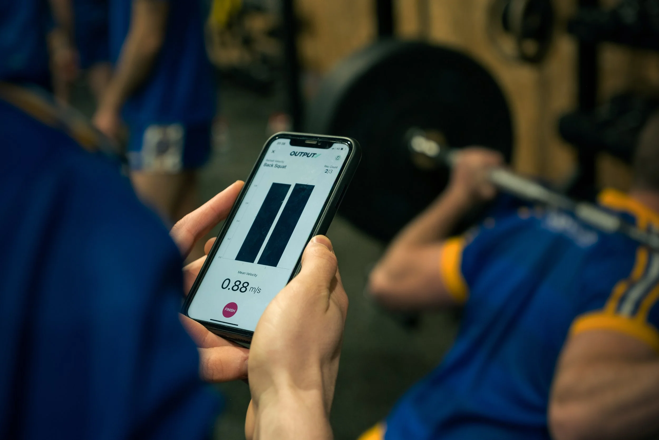 A person holding a smartphone displaying a fitness app with a bar graph and the text 'Back Squat', along with a speed measurement of 0.88 meters per second. In the background, other people are seen, some wearing sportswear, and gym equipment is visible.