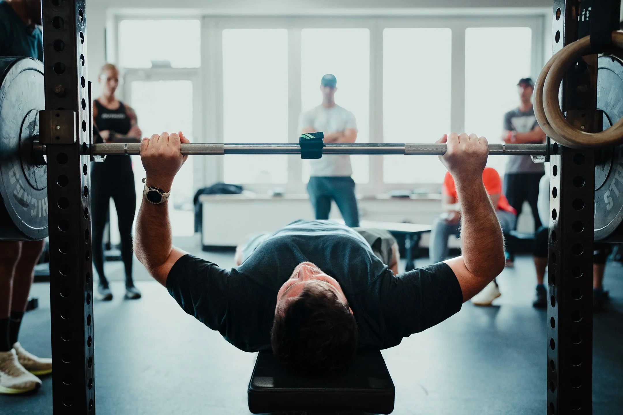 Person lying on a bench pressing weights in a gym, with people watching in the background.