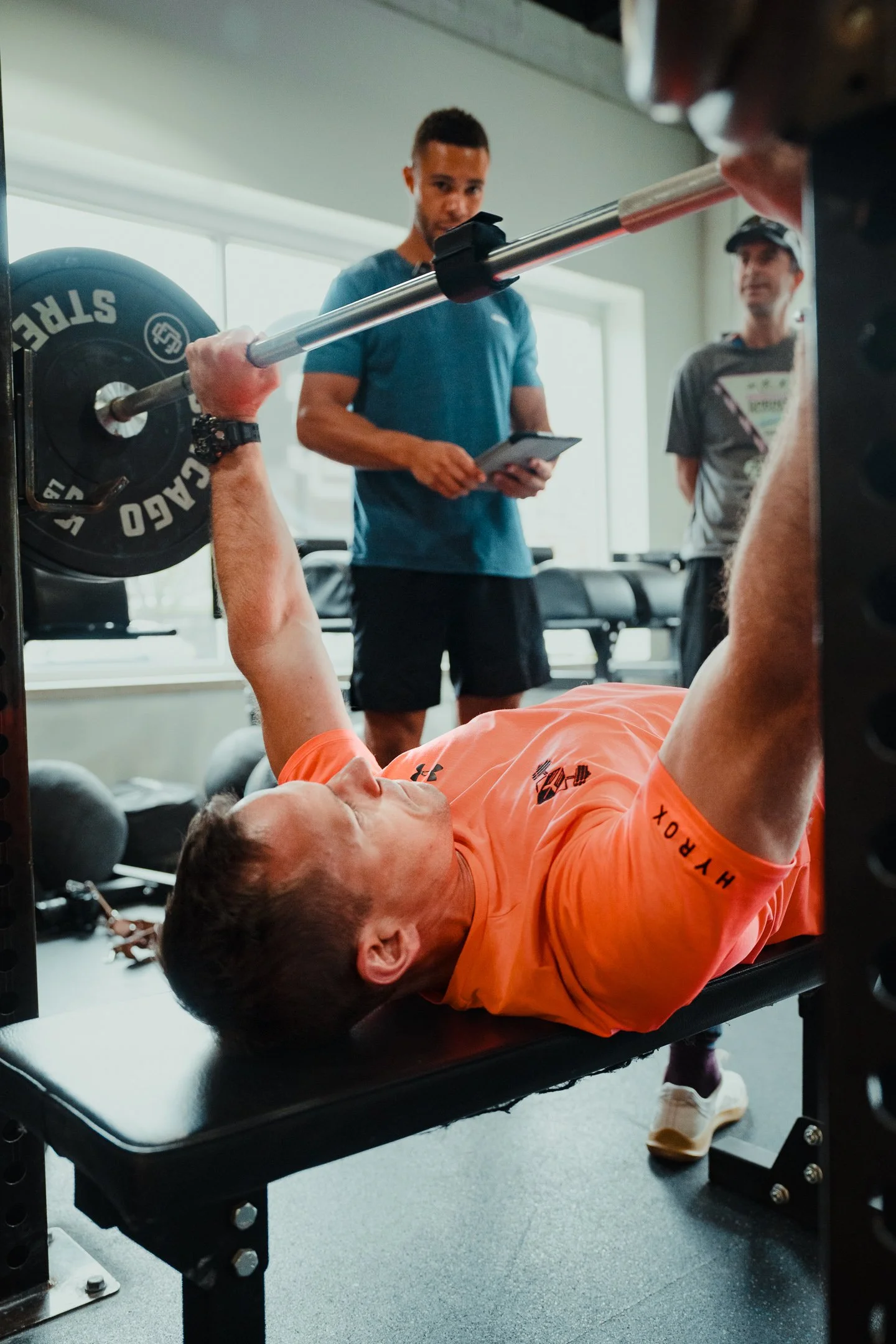 A person in an orange shirt lying on a workout bench lifting a barbell with weights, while two other individuals observe, one of whom is holding a tablet. The gym has large windows and various weights and equipment visible.