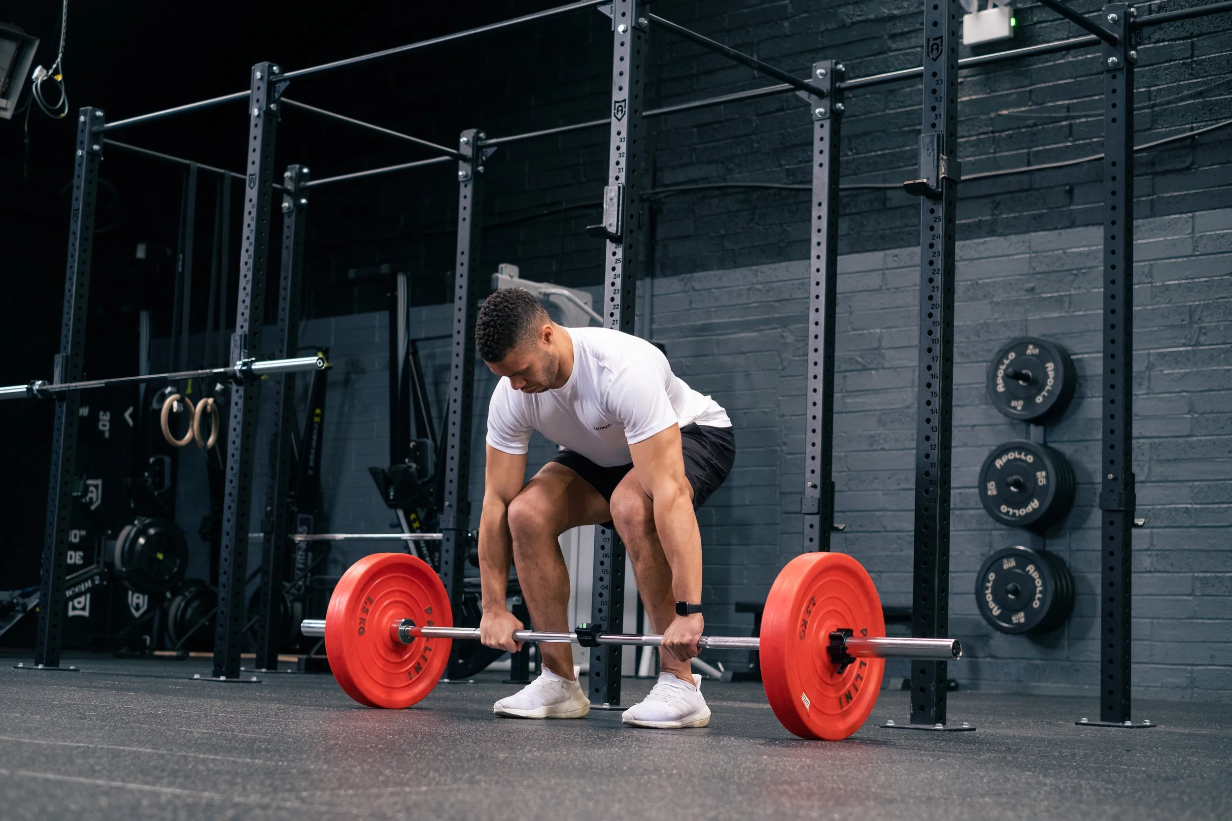 A man in a white t-shirt and black shorts is lifting a barbell with red weight plates in a gym, with black brick walls and gym equipment in the background.