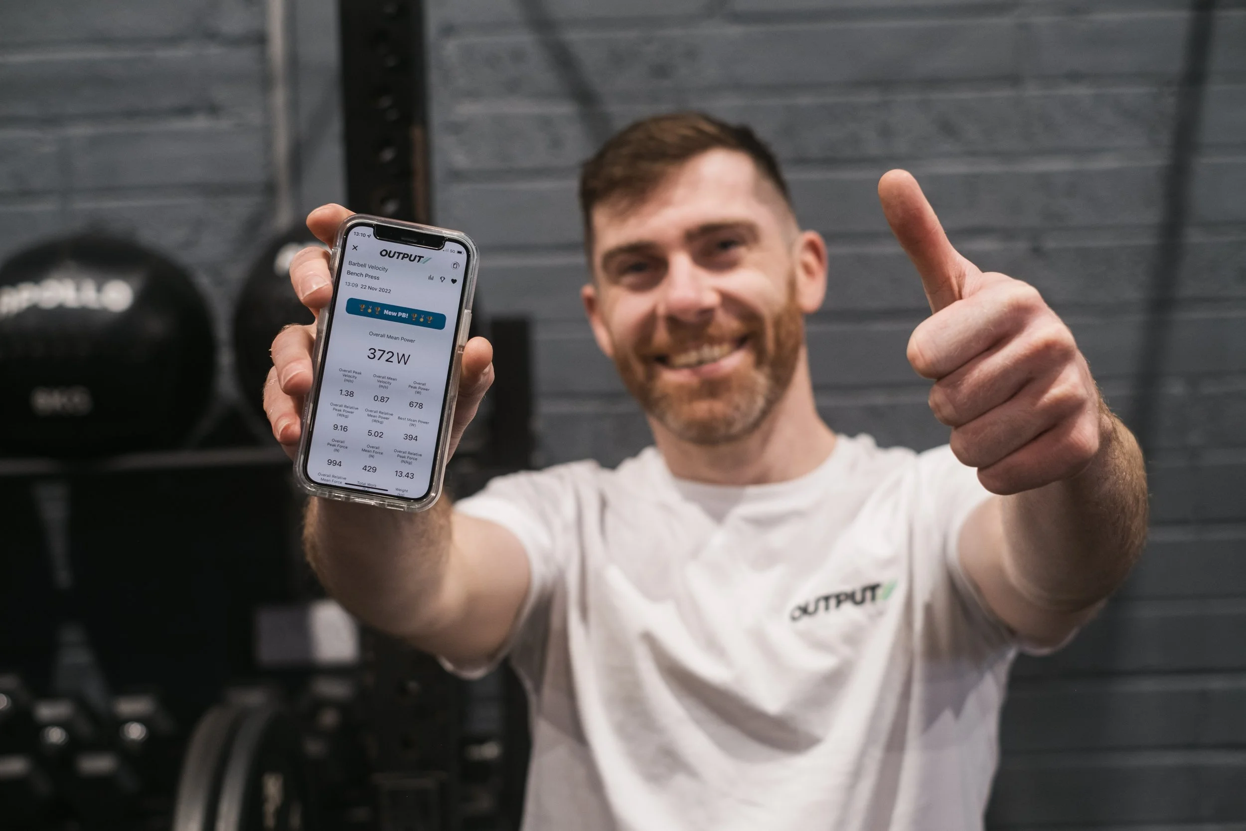 A smiling man holding a smartphone displaying workout data while giving a thumbs-up in a gym setting.