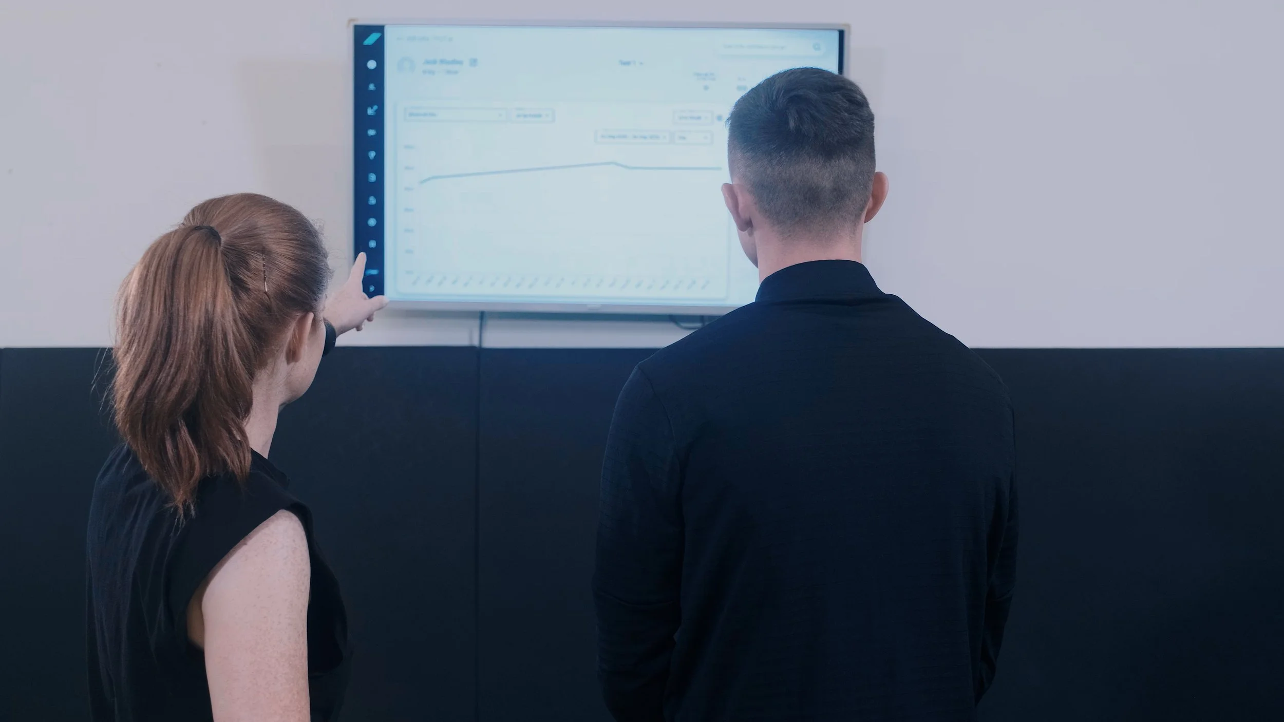 Two people, a woman with red hair and a man with dark hair, stand in front of a wall-mounted digital display showing a line graph and data. The woman is pointing at the screen.