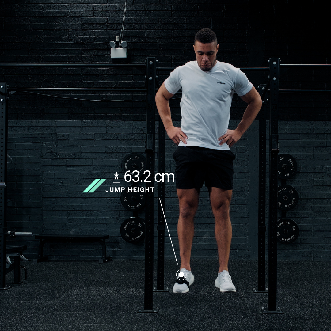 A man stands with his hands on his hips in a gym, preparing to jump. A graphic overlay shows his jump height as 63.2 cm.