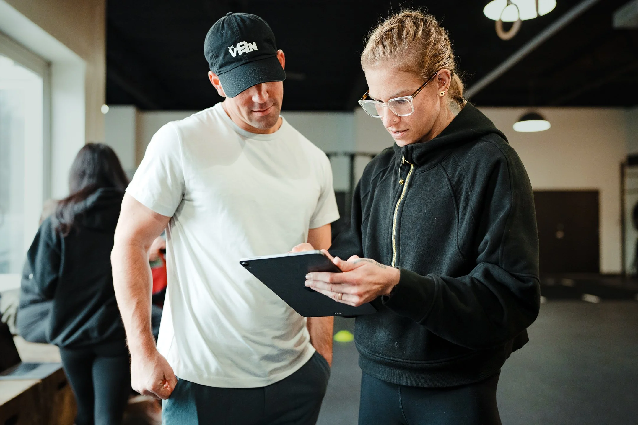 A man and woman looking at a tablet together indoors with gym equipment visible in the background.