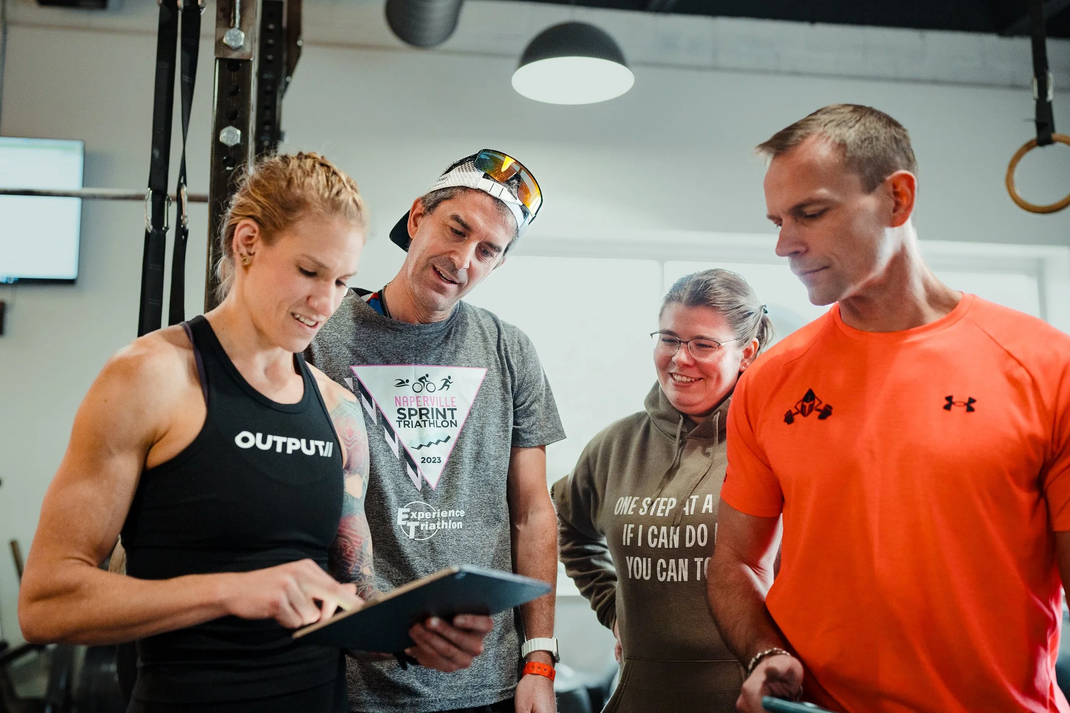 Group of five people looking at a tablet, two men and three women, some wearing workout attire, in a gym or fitness setting.