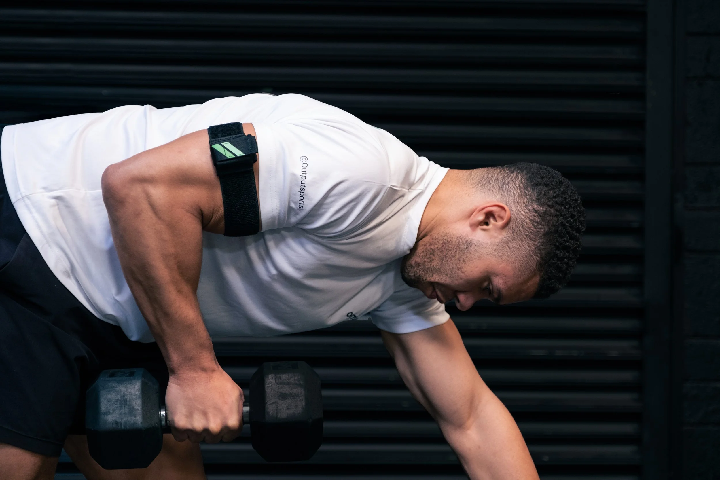 A man with short, curly hair, wearing a white T-shirt and black shorts, is leaning forward working out with a dumbbell in a gym. The background features black horizontal lines.