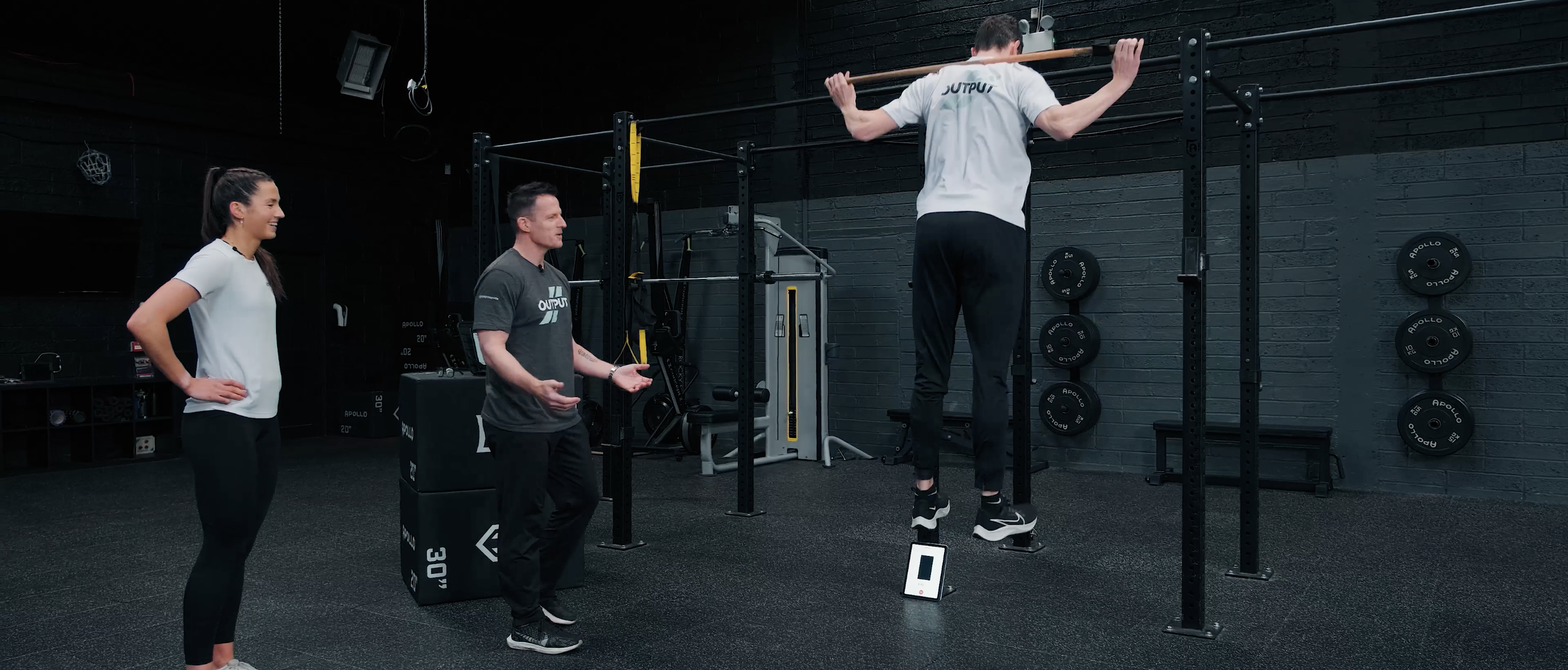 Person doing pull-ups on gym equipment while two trainers or observers watch in a black-walled gym.