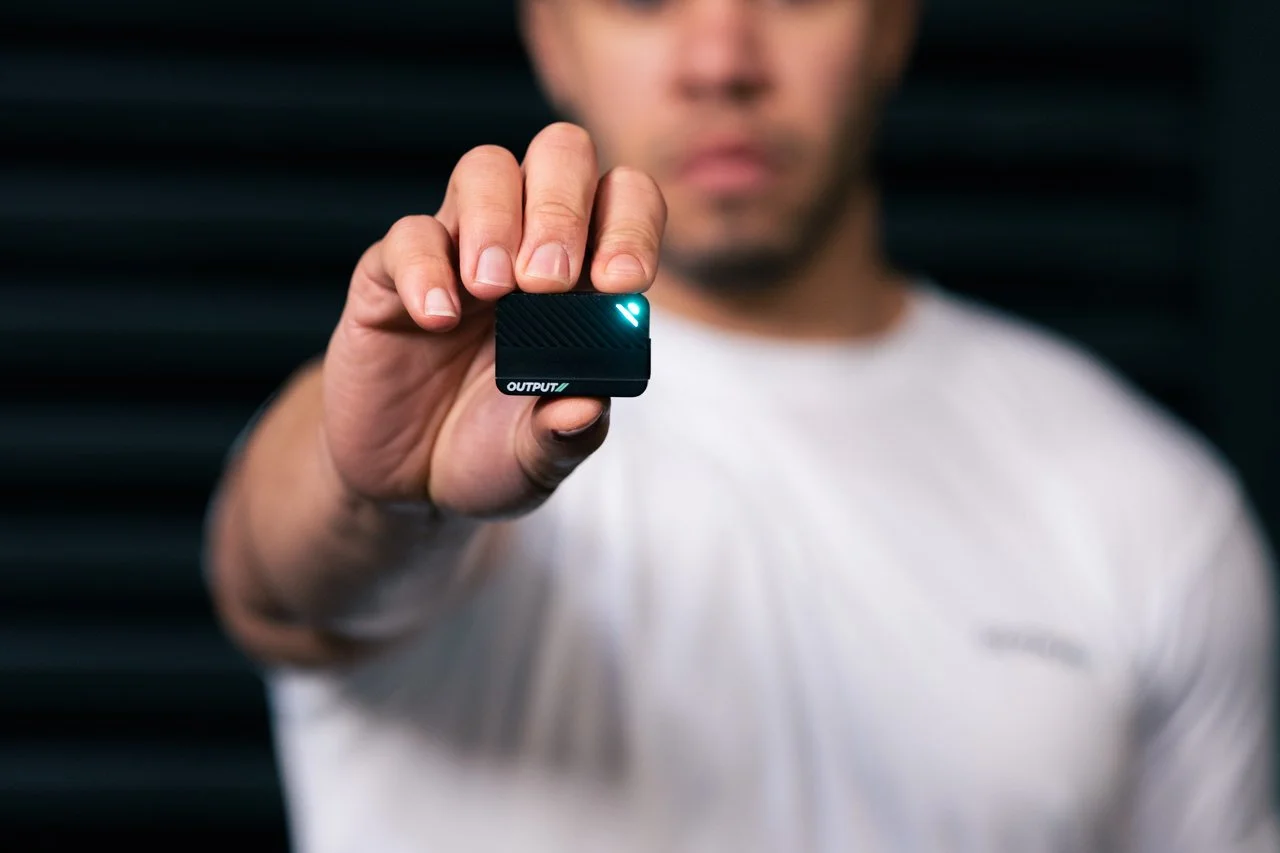 Person holding a small black electronic device with a glowing blue light and the word 'OUTPUT' visible on it, with a blurred background.
