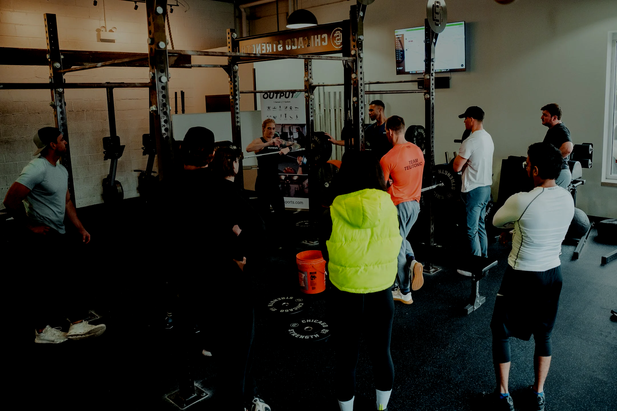 A group of people in a gym participating in a fitness class or training session, standing around a squat rack with weights and barbells, watching an instructor demonstrate or explain something.
