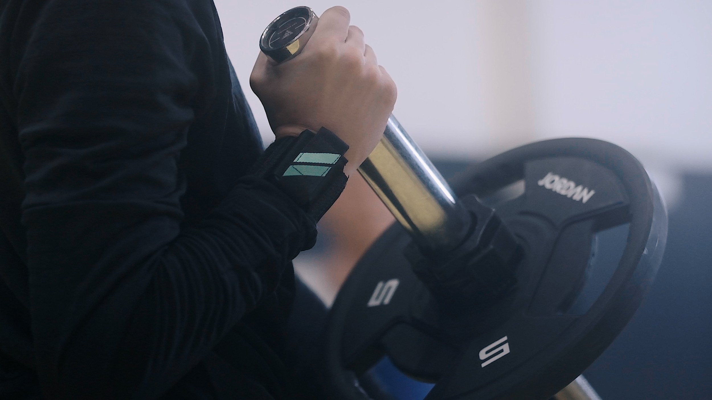 A person in a black jacket holding a gold and black dumbbell, alongside a black weight plate with white writing, in a gym.