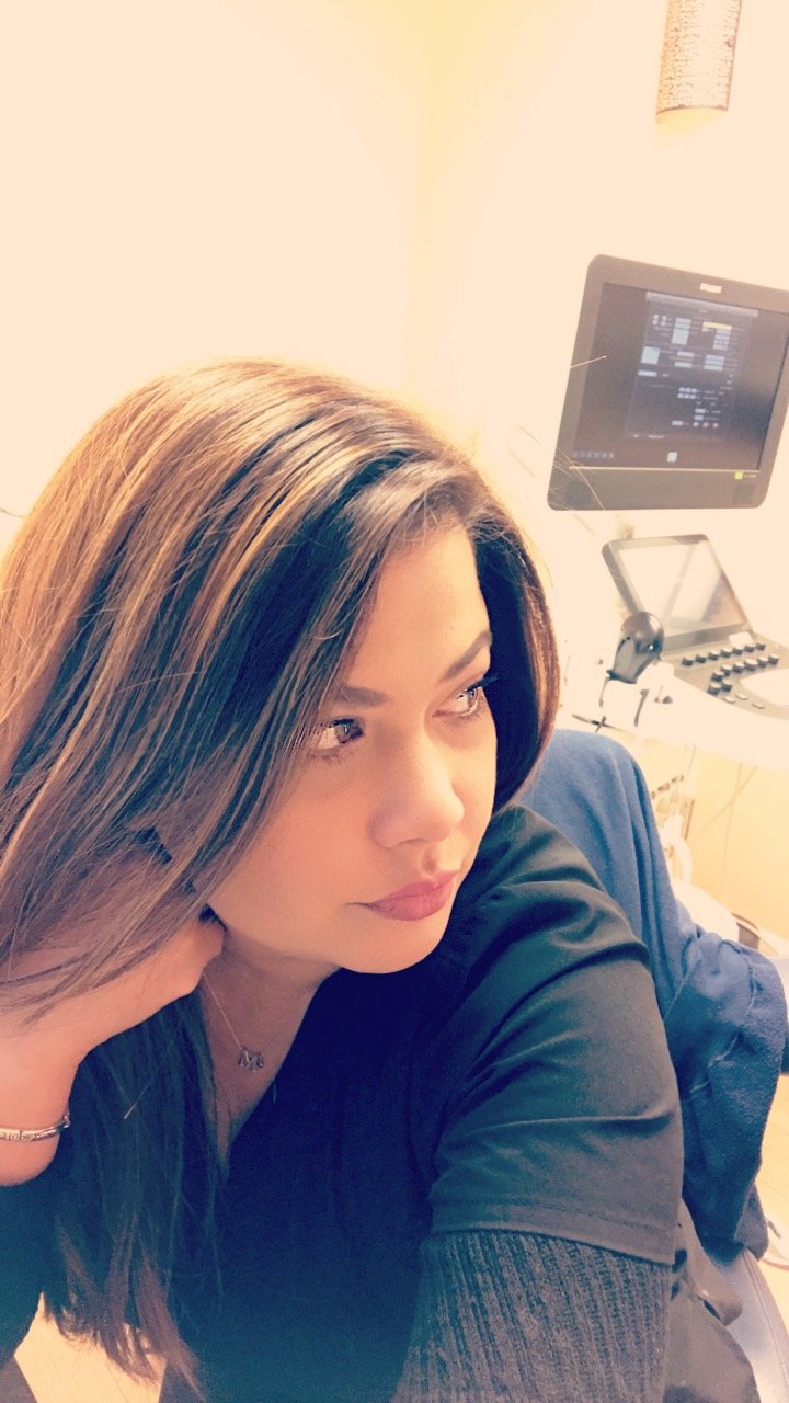 A woman with long, brown hair resting her head on her hand, lying in a hospital bed with medical monitors visible in the background.
