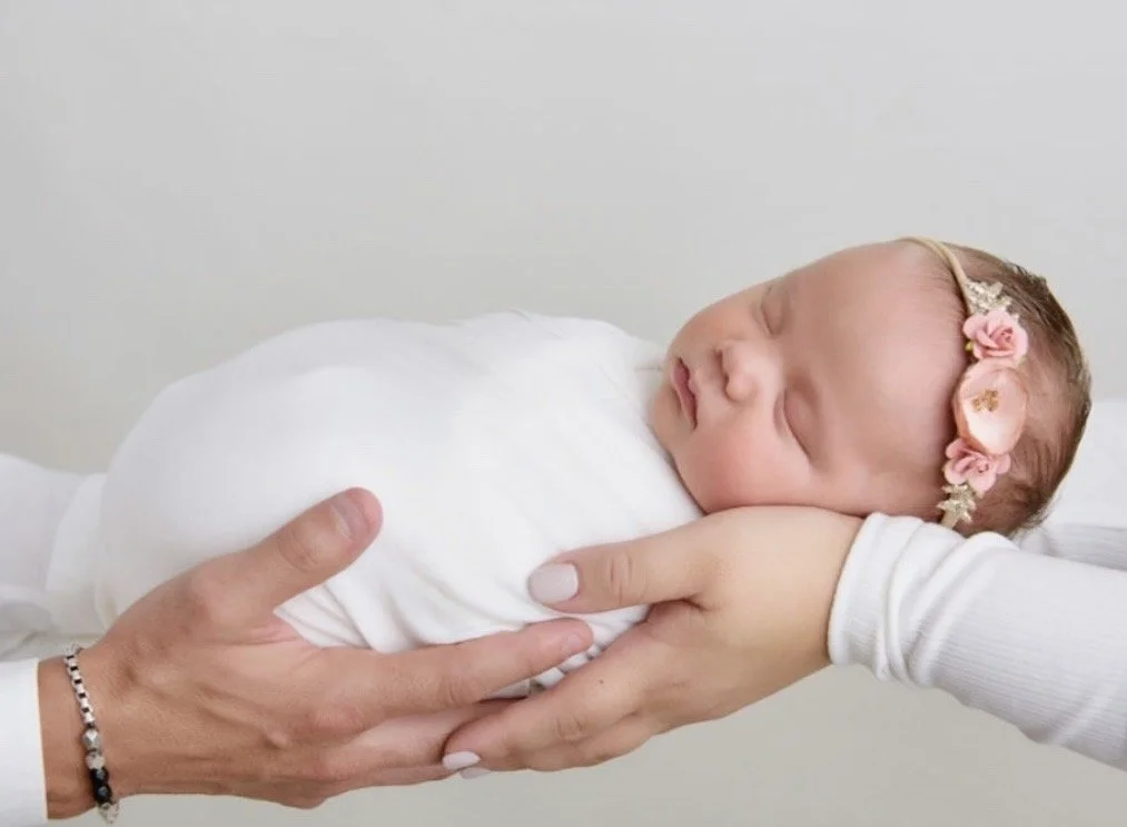 A sleeping newborn baby girl with a floral headband being gently cradled in adult hands, against a plain white background.