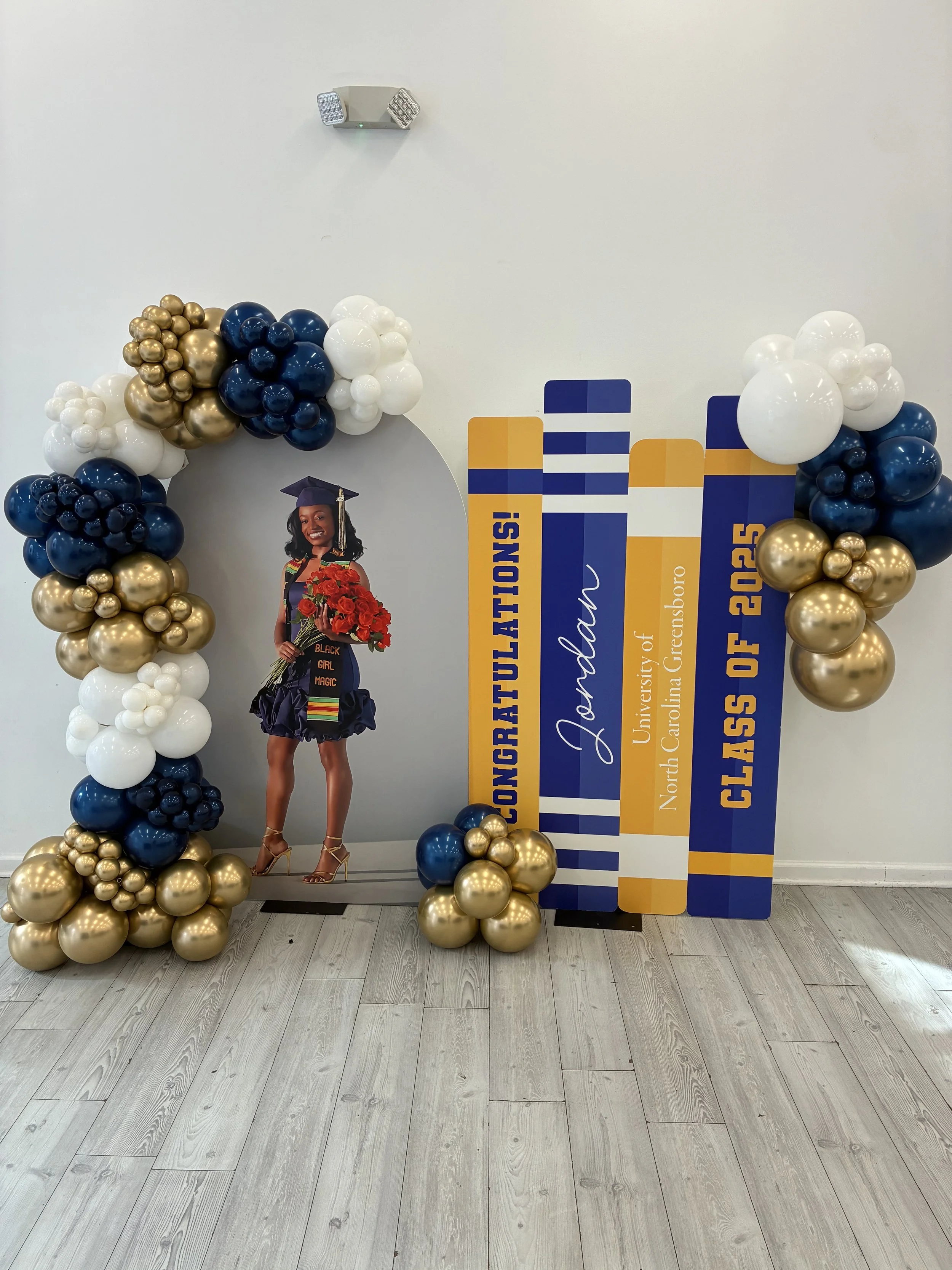 Decorated photo backdrop with balloons in white, blue, and gold, a cutout of a young woman in a graduation cap and gown holding a bouquet of red roses, and congratulatory text for the class of 2025 graduation from North Carolina Greensboro, with the name Jared.