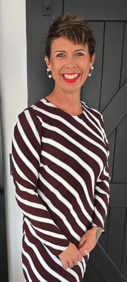 Woman smiling with short dark hair, wearing pearl earrings, red lipstick, and a long-sleeved dress with white and dark maroon diagonal stripes, standing in front of a black door.