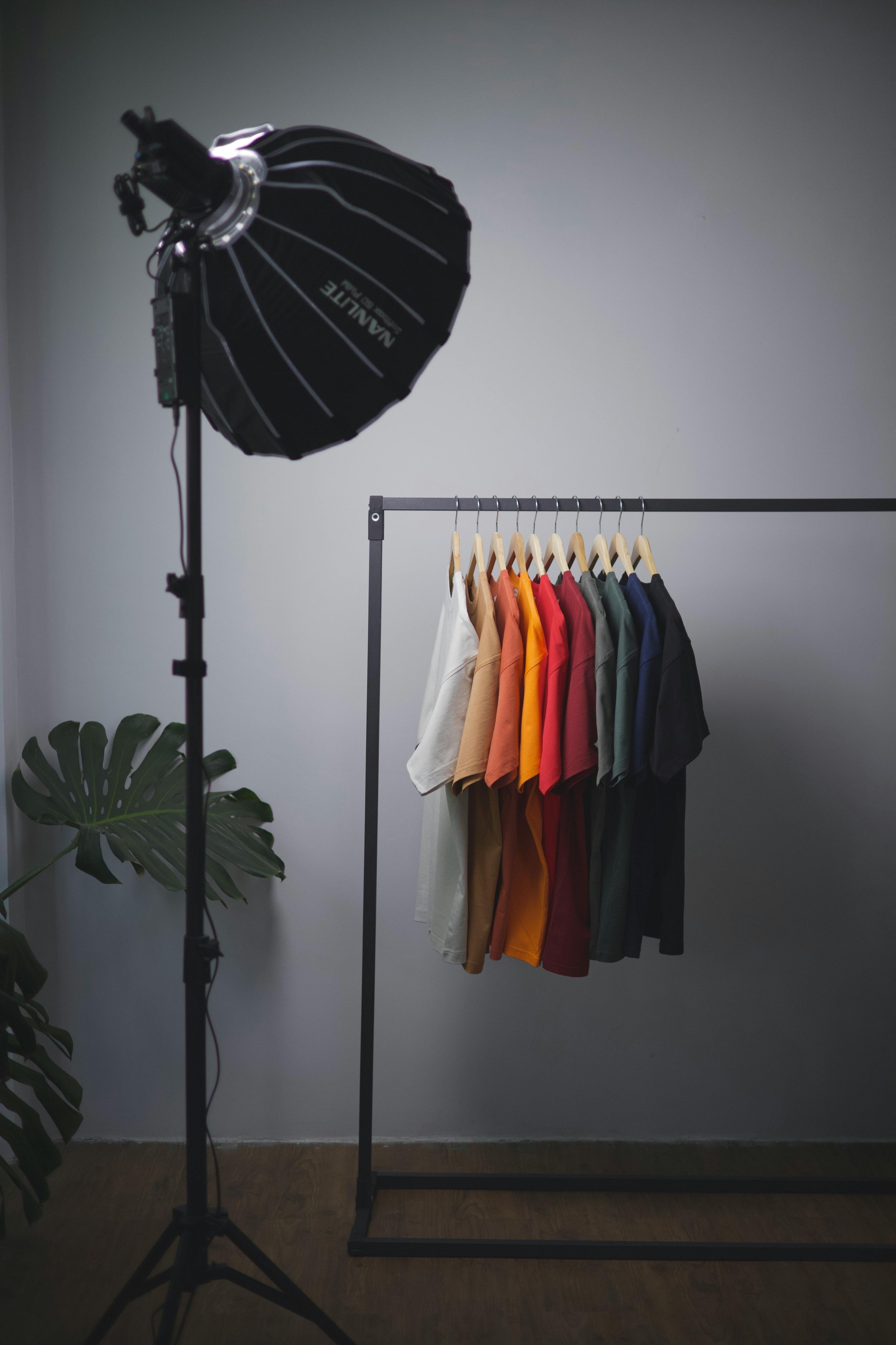 Photo of a clothing rack with multicolored T-shirts in front of a plain wall. A large studio light is positioned to the left, and a green plant is partially visible on the bottom left.