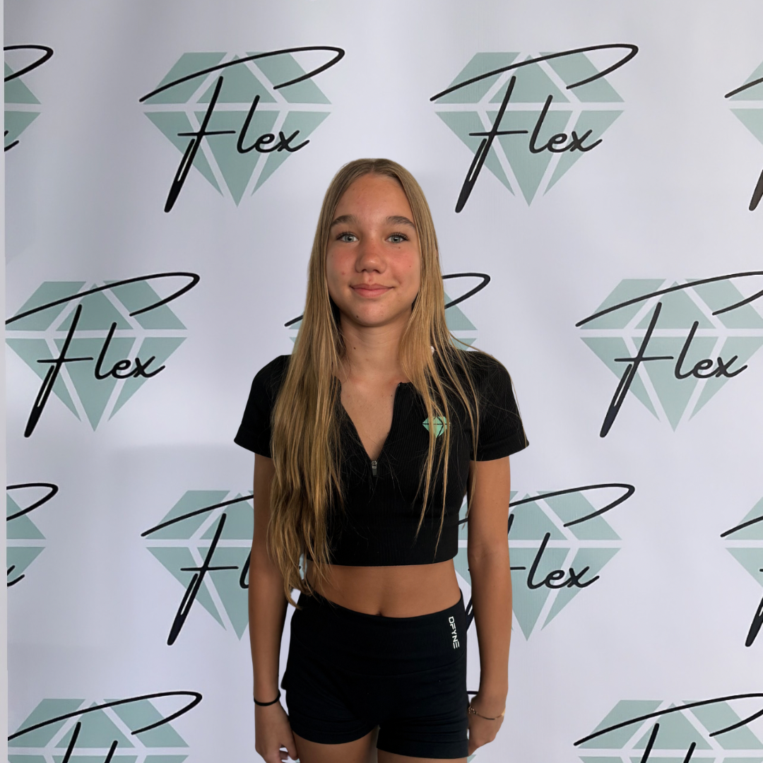A teenage girl with long blonde hair standing in front of a backdrop with a diamond logo and the word 'Flex' in black cursive writing. She is wearing a black crop top and black athletic shorts, with a slight smile.