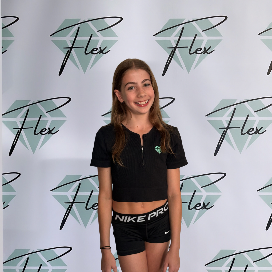 A young girl with long brown hair, smiling, standing in front of a backdrop with the Flex logo, wearing a black crop top with a small green diamond logo and black Nike Pro shorts.