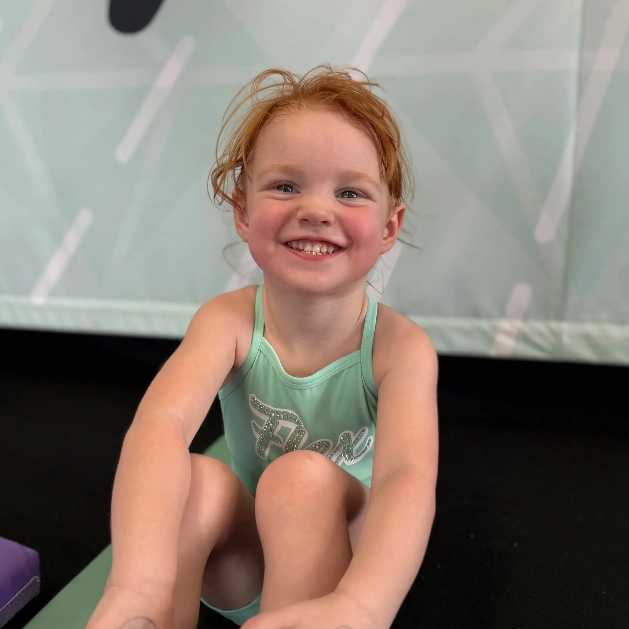 A smiling young girl with red hair and fair skin sitting on a black surface, wearing a mint green tank top with decorative lettering, in front of a light green backdrop with white streaks.