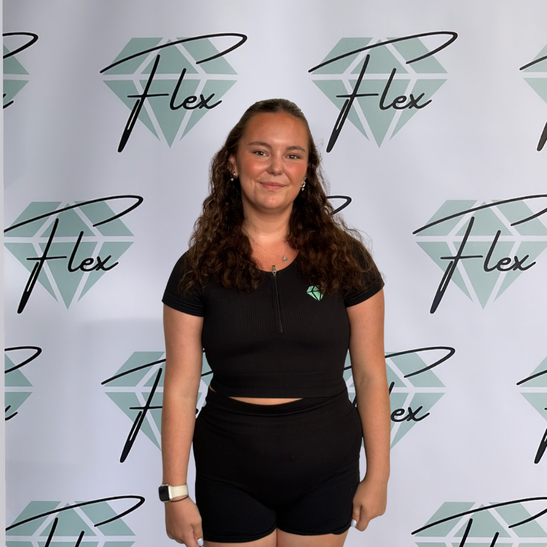 A woman standing in front of a backdrop with a diamond logo and the word 'Flex' repeated. She has curly brown hair, is smiling, and is dressed in a black athletic outfit with a logo on her top.
