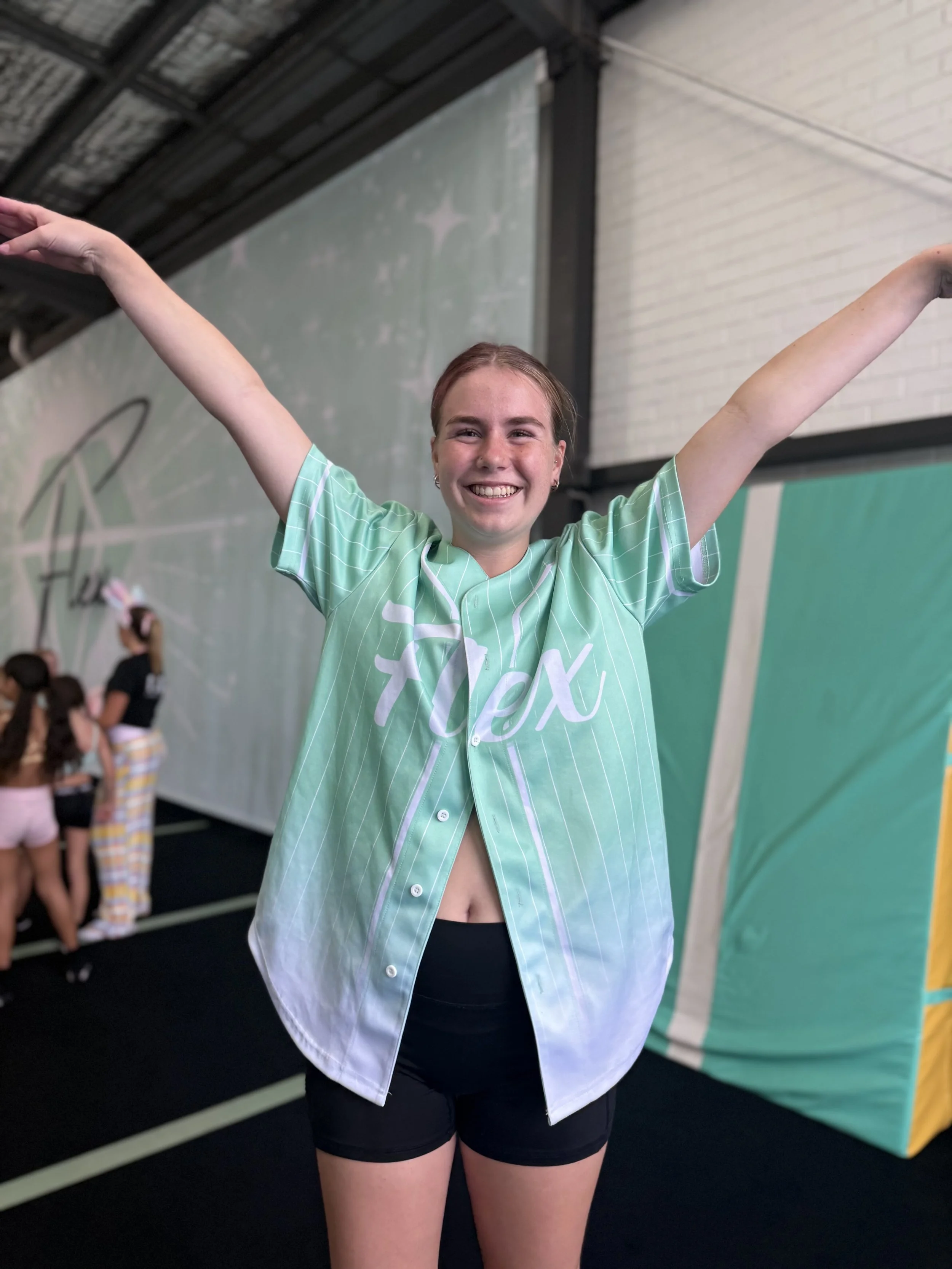 A young female gymnast smiling with arms raised in a gymnasium with other children in the background.