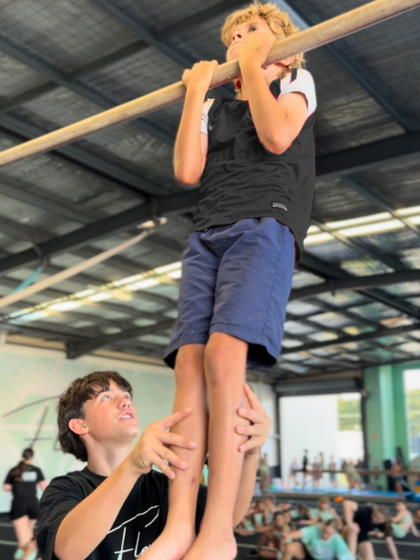 A boy practicing a pull-up exercise on a wooden bar in a gym with other people exercising in the background.