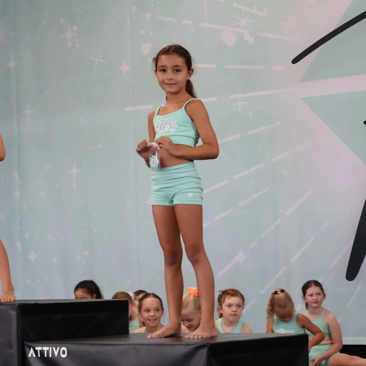 Young girl in matching light blue dance exercise outfit standing on winners' podium at a competition, holding a medal, with other girls sitting behind her.