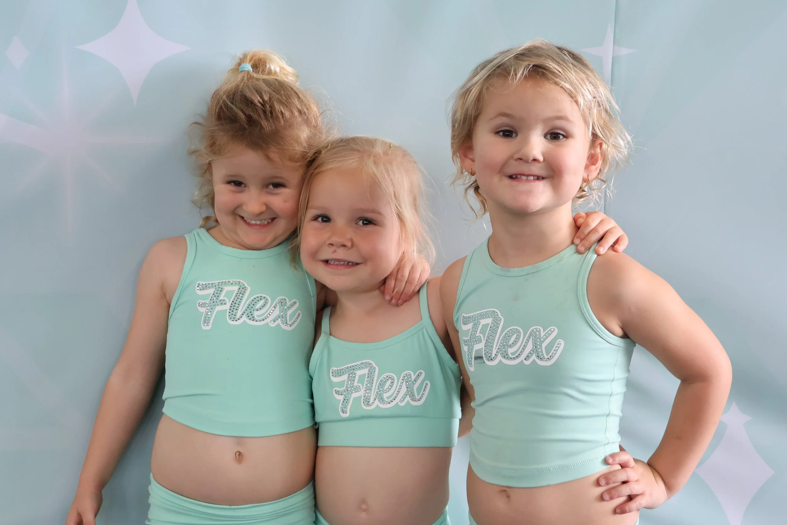 Three young girls in matching turquoise cheerleader outfits with the word 'Flex' written on the front, smiling and posing together against a light blue background with star-like designs.