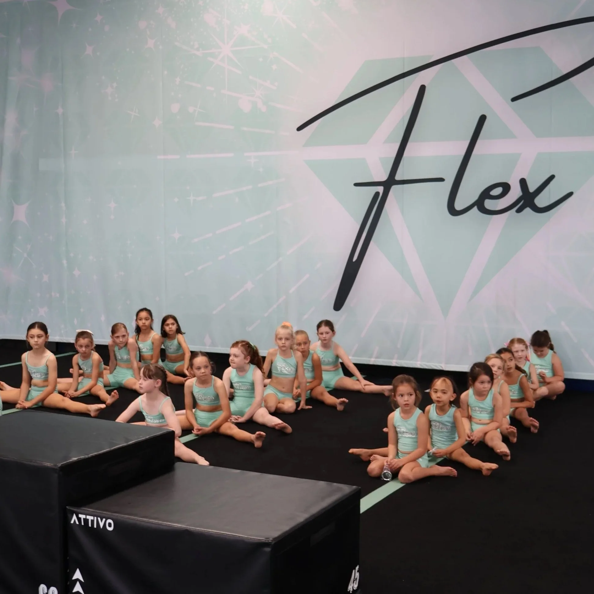 Young girls in turquoise dance costumes sitting on the floor of a gym or dance studio, with a large backdrop featuring the word 'Flex' and a diamond logo behind them.